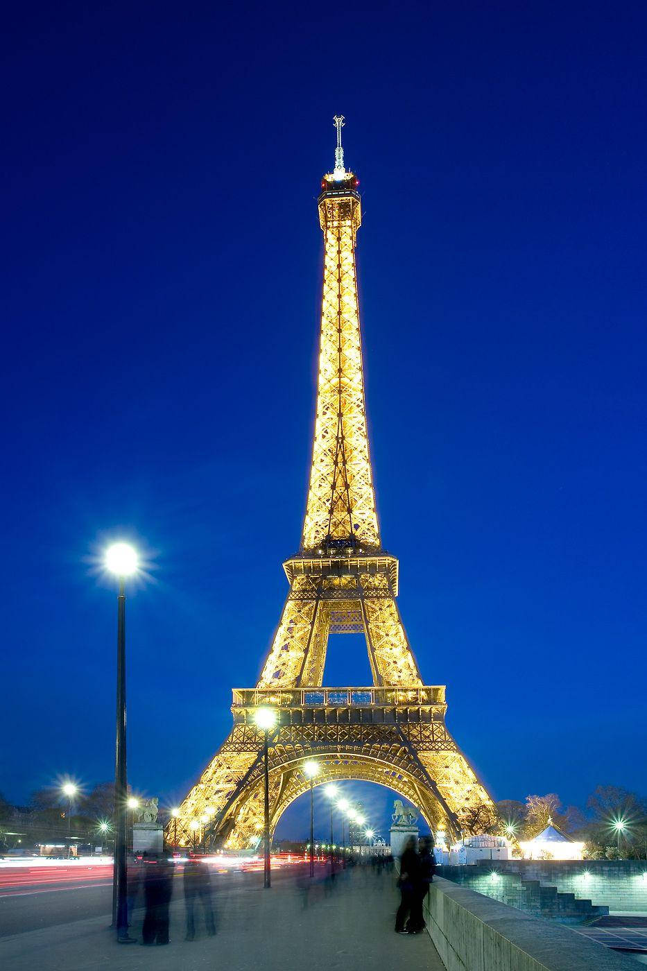 Paris At Night 桌布 壁紙 — 艾菲爾鐵塔在深藍色的暮色天空中閃耀著金色光芒，城市燈光形成模糊的運動軌跡。