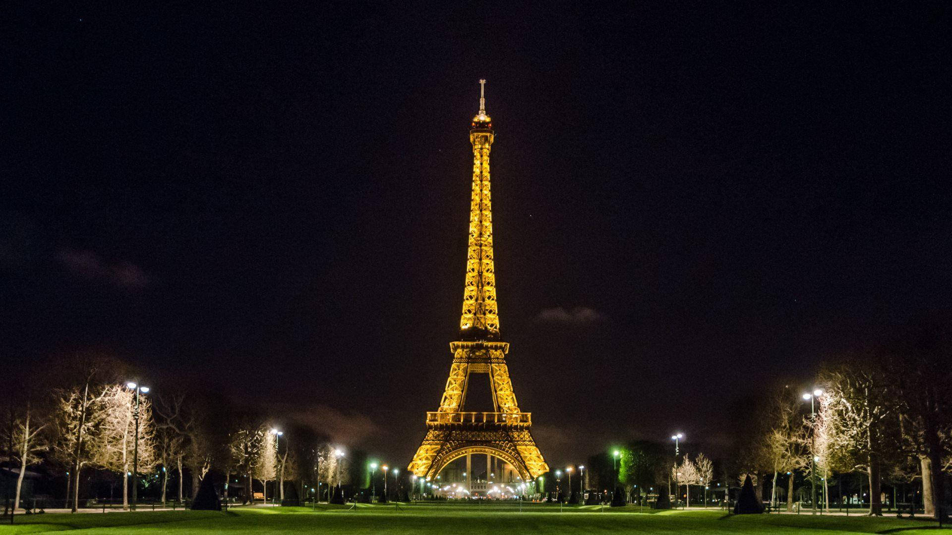 Paris Eiffel Tower 桌布 壁紙 — 艾菲爾鐵塔在深邃的夜空下溫暖地發光，被光禿的樹木和公園燈光框住，呈現寧靜的巴黎場景。