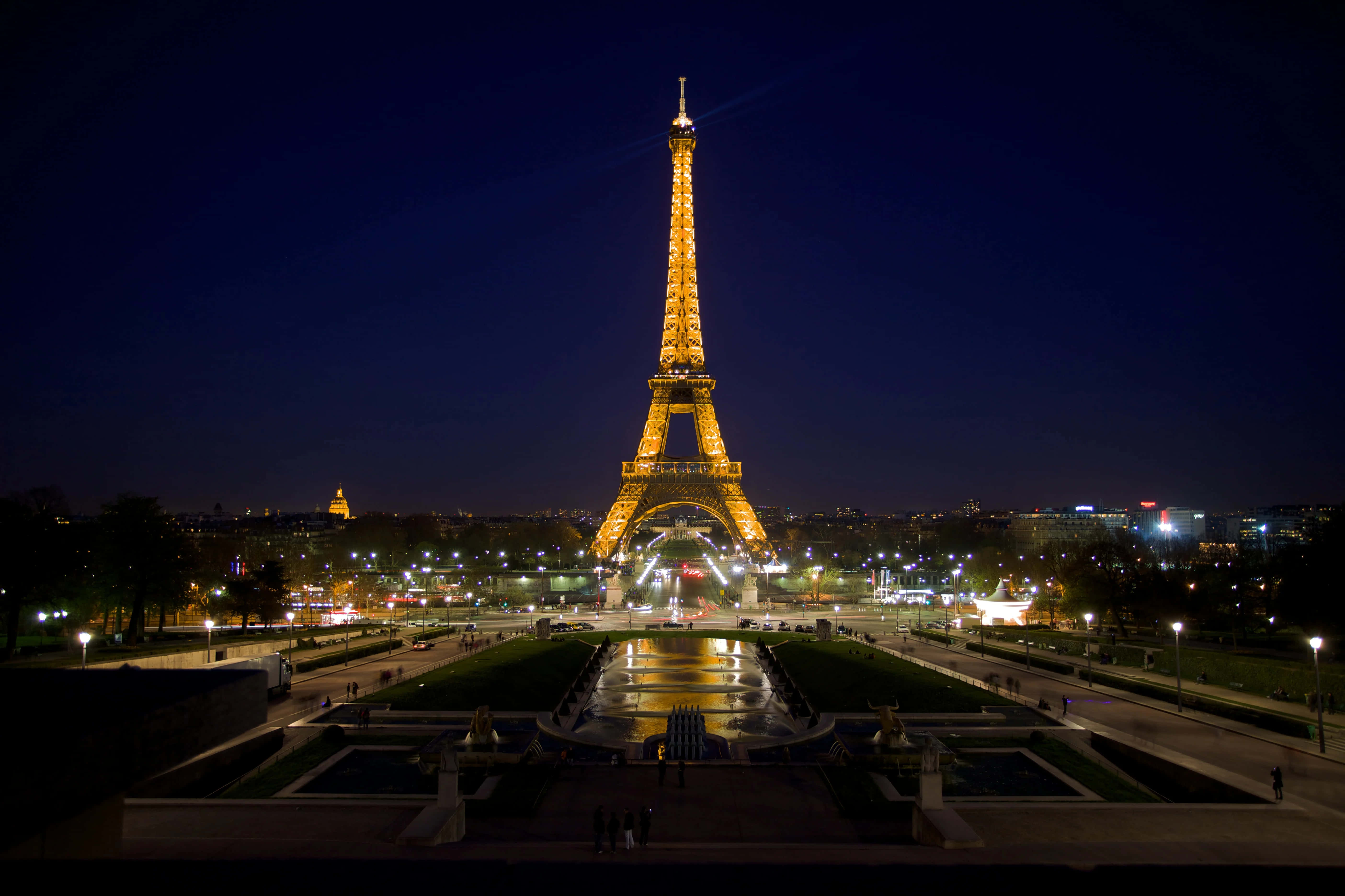 Eiffel Tower 桌布 壁紙 — 艾菲爾鐵塔在深邃的夜空中溫暖地發光，其燈光倒映在中央噴泉中，遠處的城市燈光閃爍。