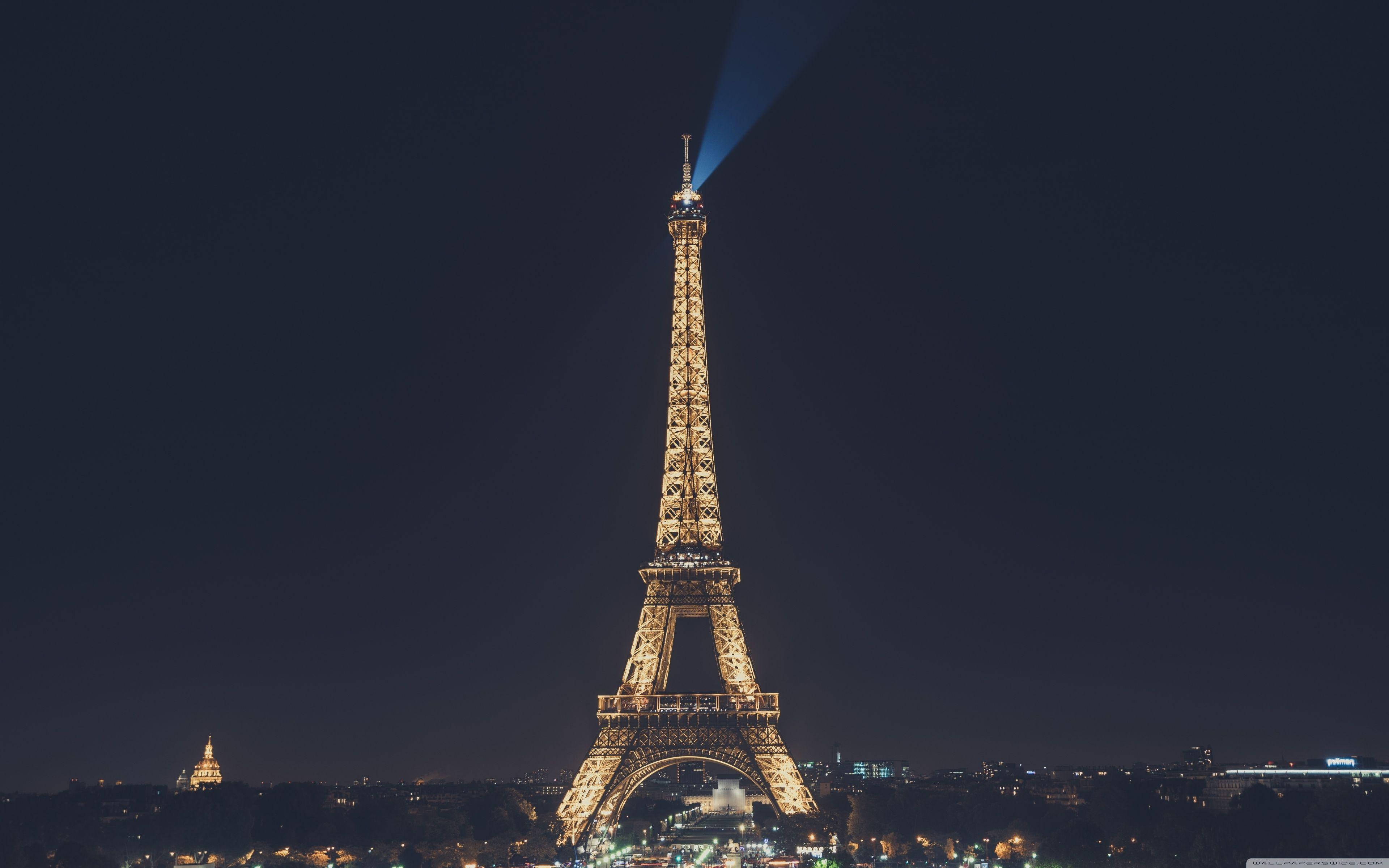 Paris Eiffel Tower 桌布 壁紙 — 艾菲爾鐵塔在深邃的夜空中溫暖地發光，一道強勁的藍色聚光燈從塔頂向上照射。