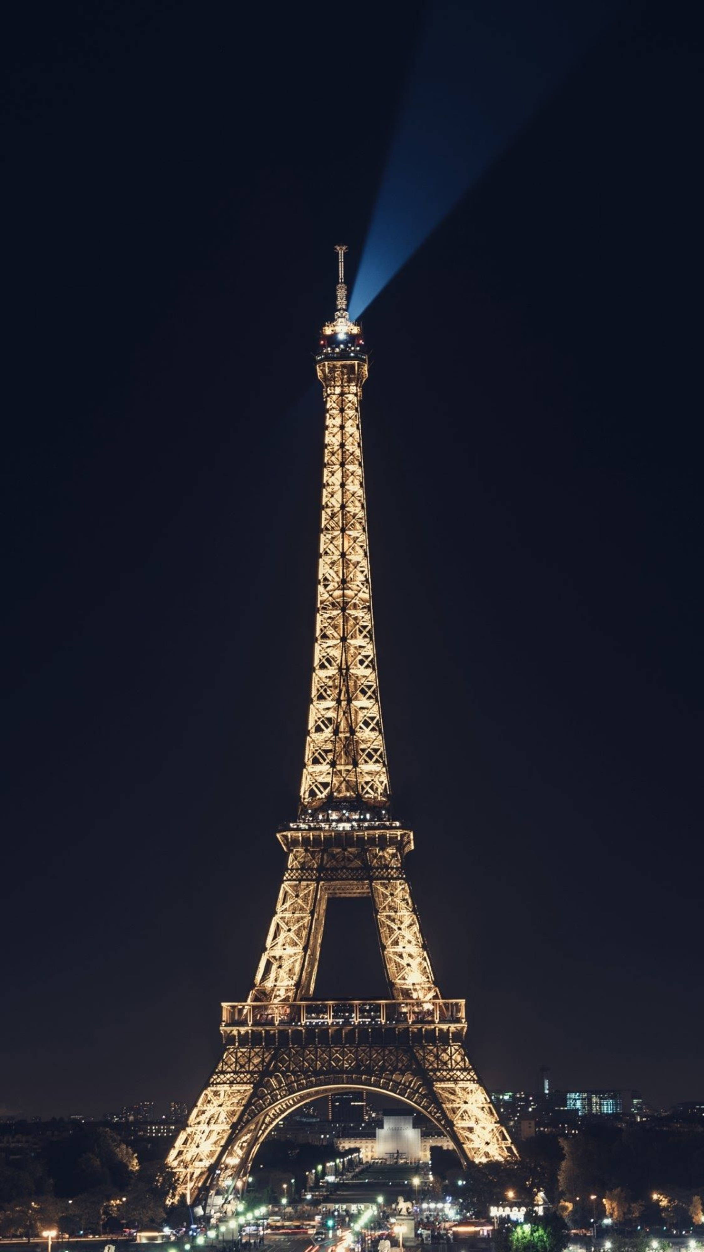 Eiffel Tower 桌布 壁紙 — 艾菲爾鐵塔在深邃的夜空中溫暖地發光，一道強勁的藍色聚光燈從其頂端向上照射。