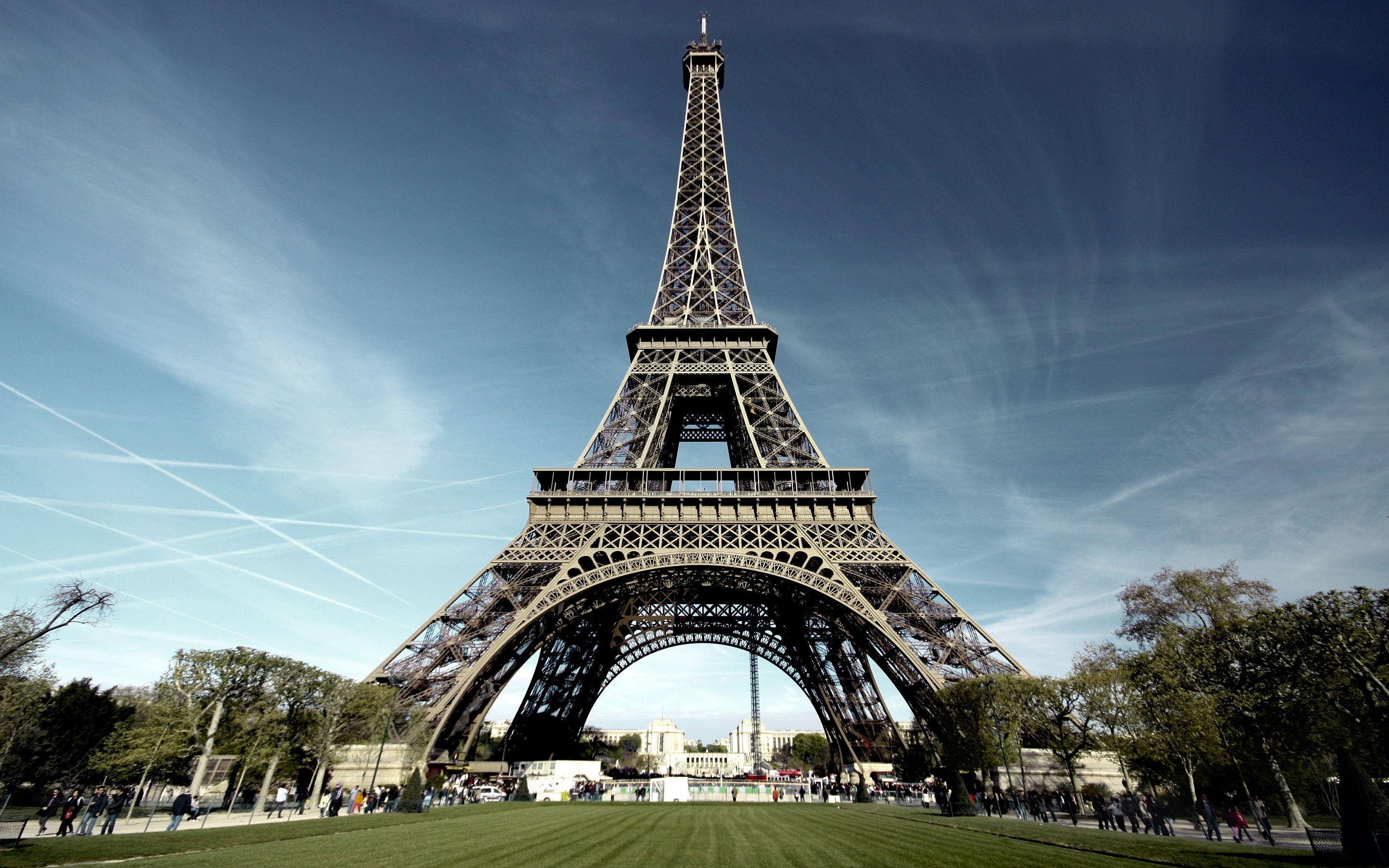 Paris Eiffel Tower 桌布 壁紙 — 艾菲爾鐵塔在清澈的藍天中高聳，天空中有縷縷薄雲和飛機凝結尾跡，周圍環繞著綠草和樹木，附近有人們在散步。