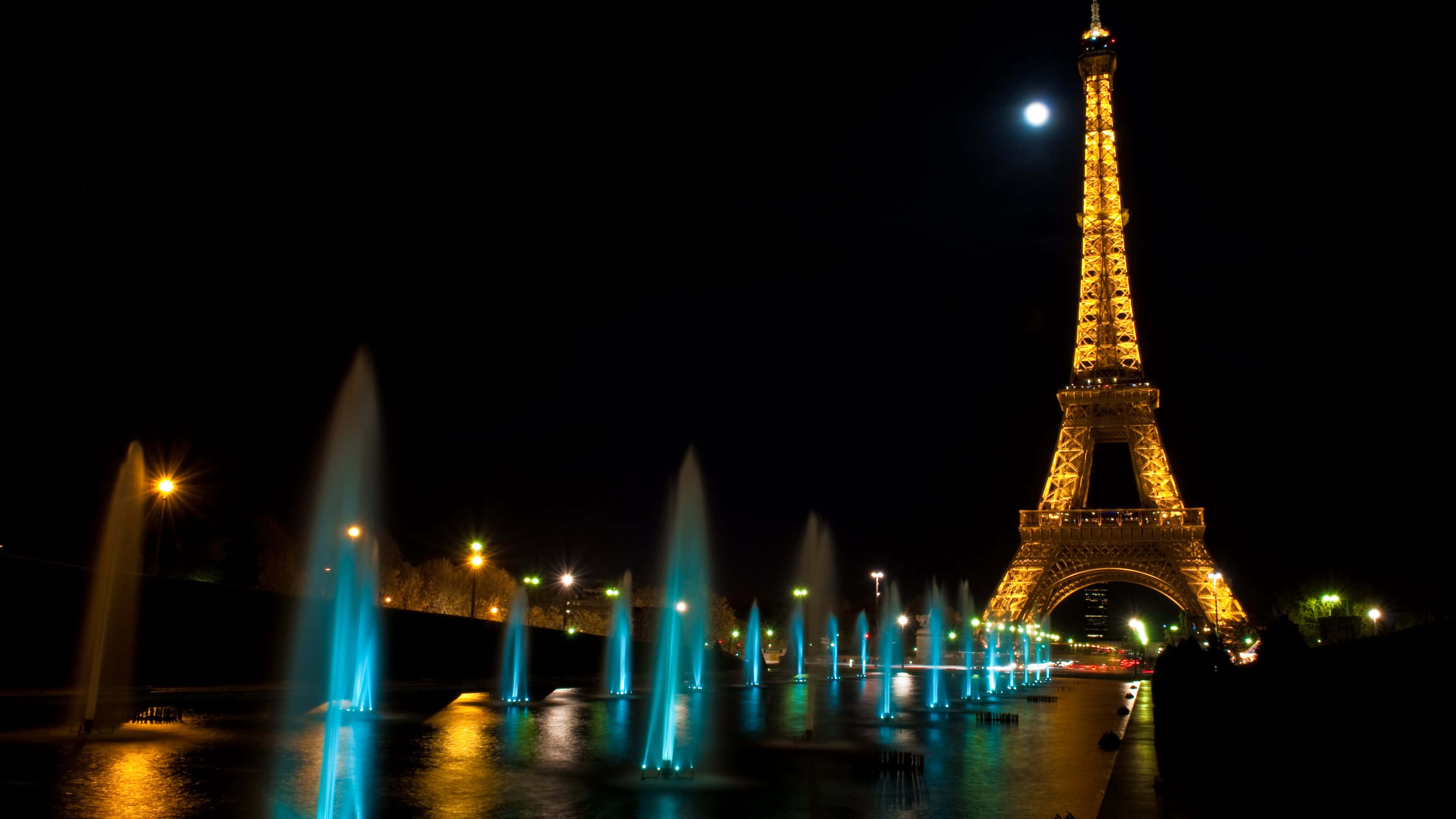 Paris At Night 桌布 壁紙 — 艾菲爾鐵塔在滿月下閃耀金光，而充滿活力的藍色噴泉在夜間前景中舞動。