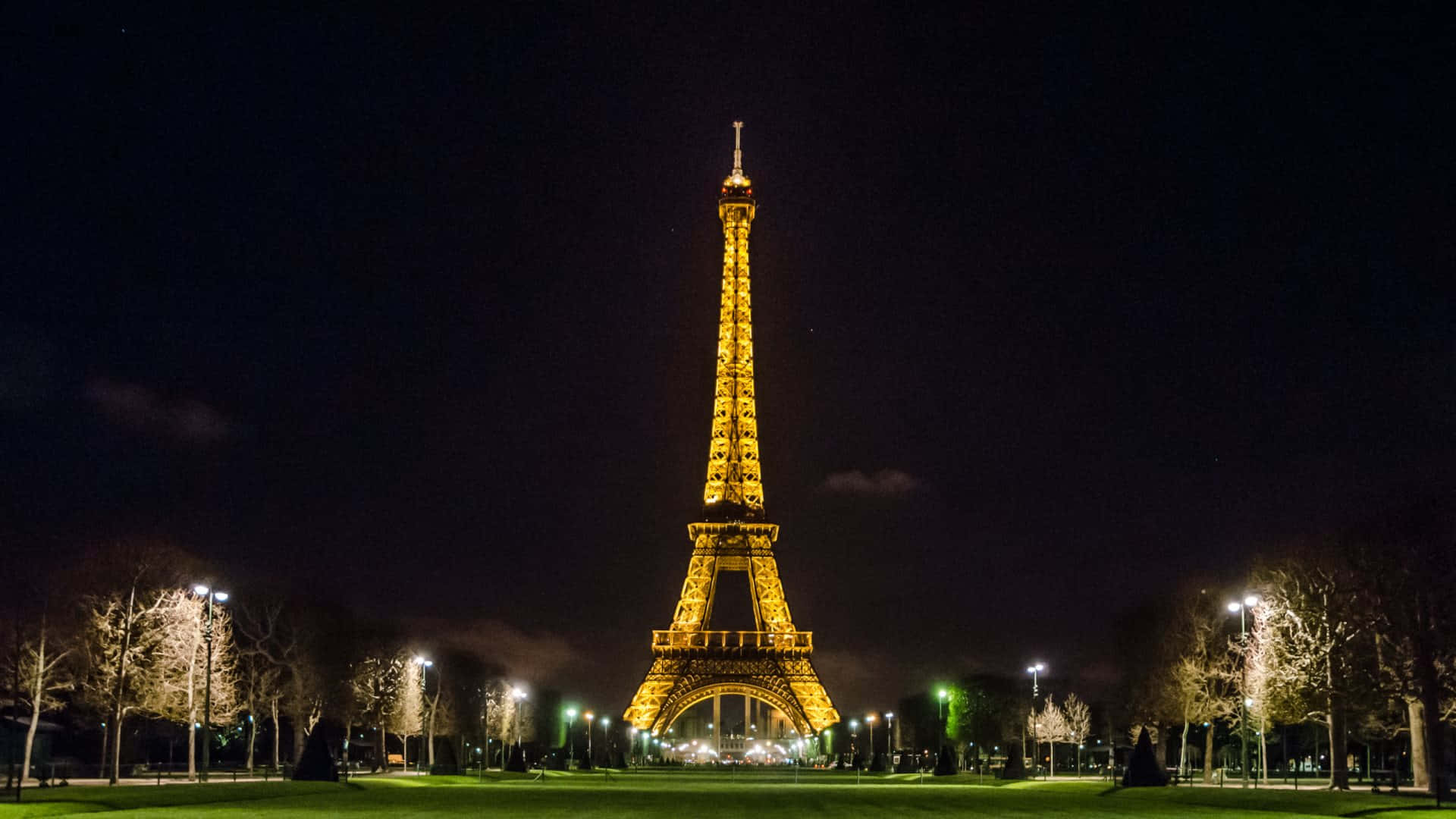 Eiffel Tower 桌布 壁紙 — 艾菲爾鐵塔在漆黑的夜空下溫暖地發光，周圍是光禿禿的樹木和柔和照亮的步道。
