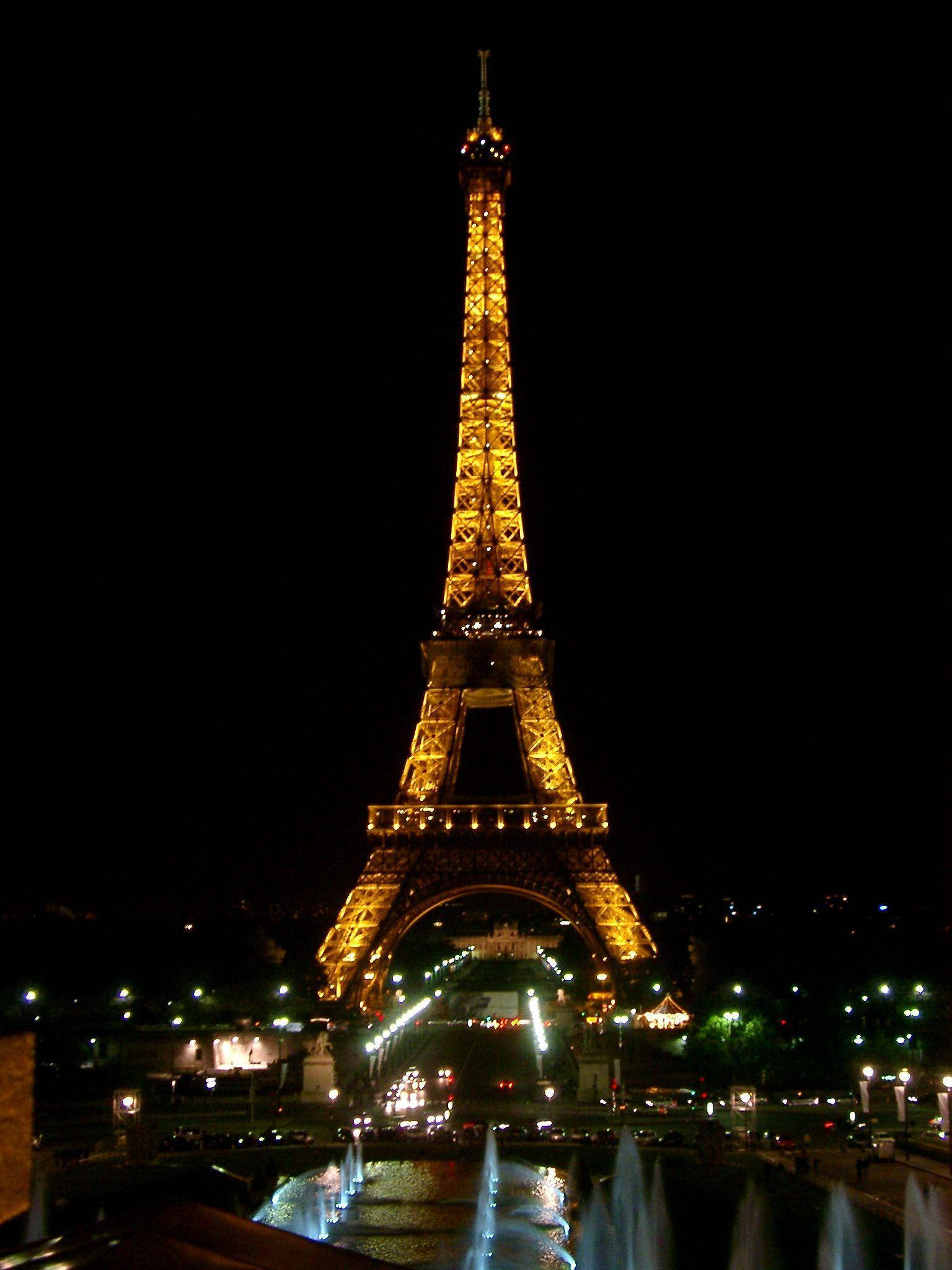 Paris Eiffel Tower 桌布 壁紙 — 艾菲爾鐵塔在漆黑的夜空下溫暖地發光，下方噴泉噴灑，遠處城市燈光閃爍。