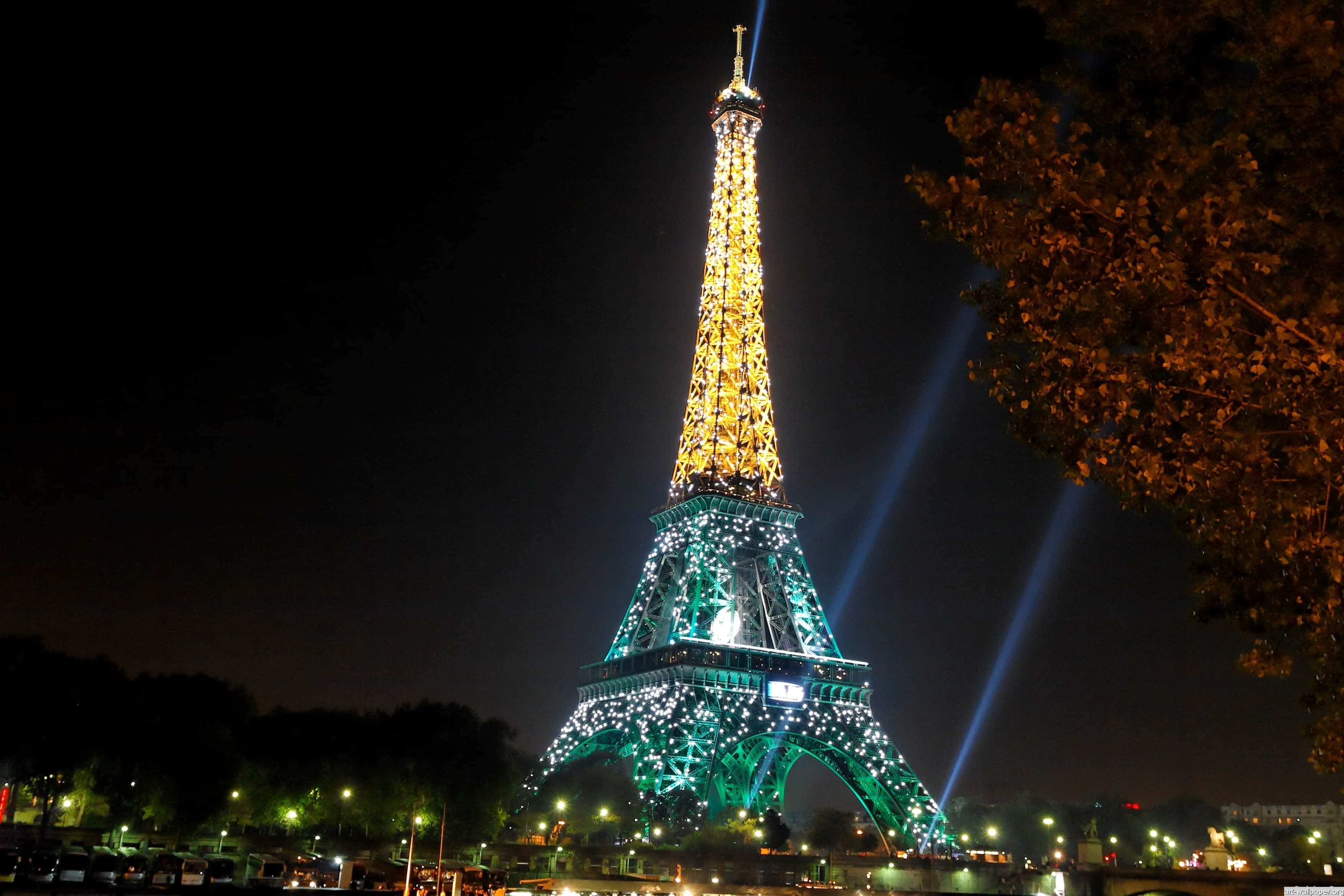 Eiffel Tower 桌布 壁紙 — 艾菲爾鐵塔在漆黑的夜空中閃耀著金色和藍綠色的燈光，光束向上射，右側有樹枝框住。