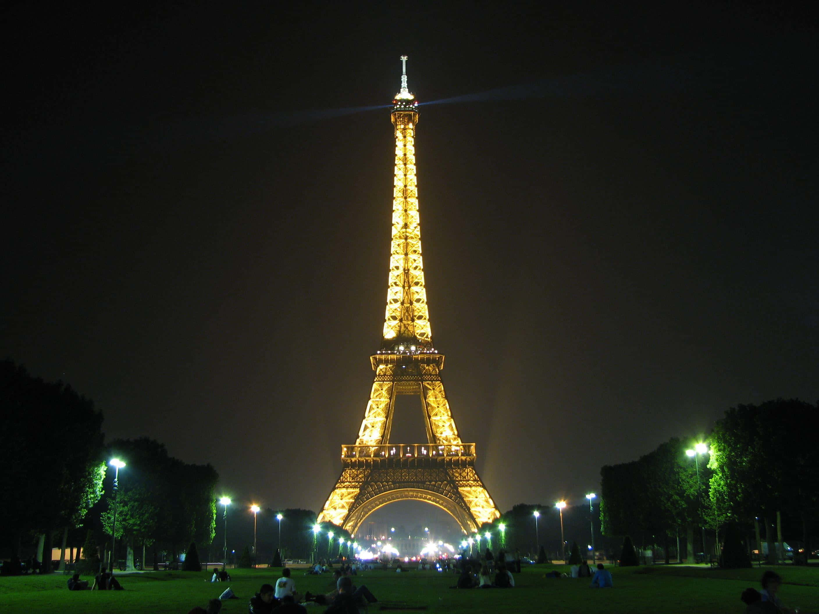 Eiffel Tower 桌布 壁紙 — 艾菲爾鐵塔在漆黑的夜空中閃耀著金色光芒，人們在下面的草地上放鬆。