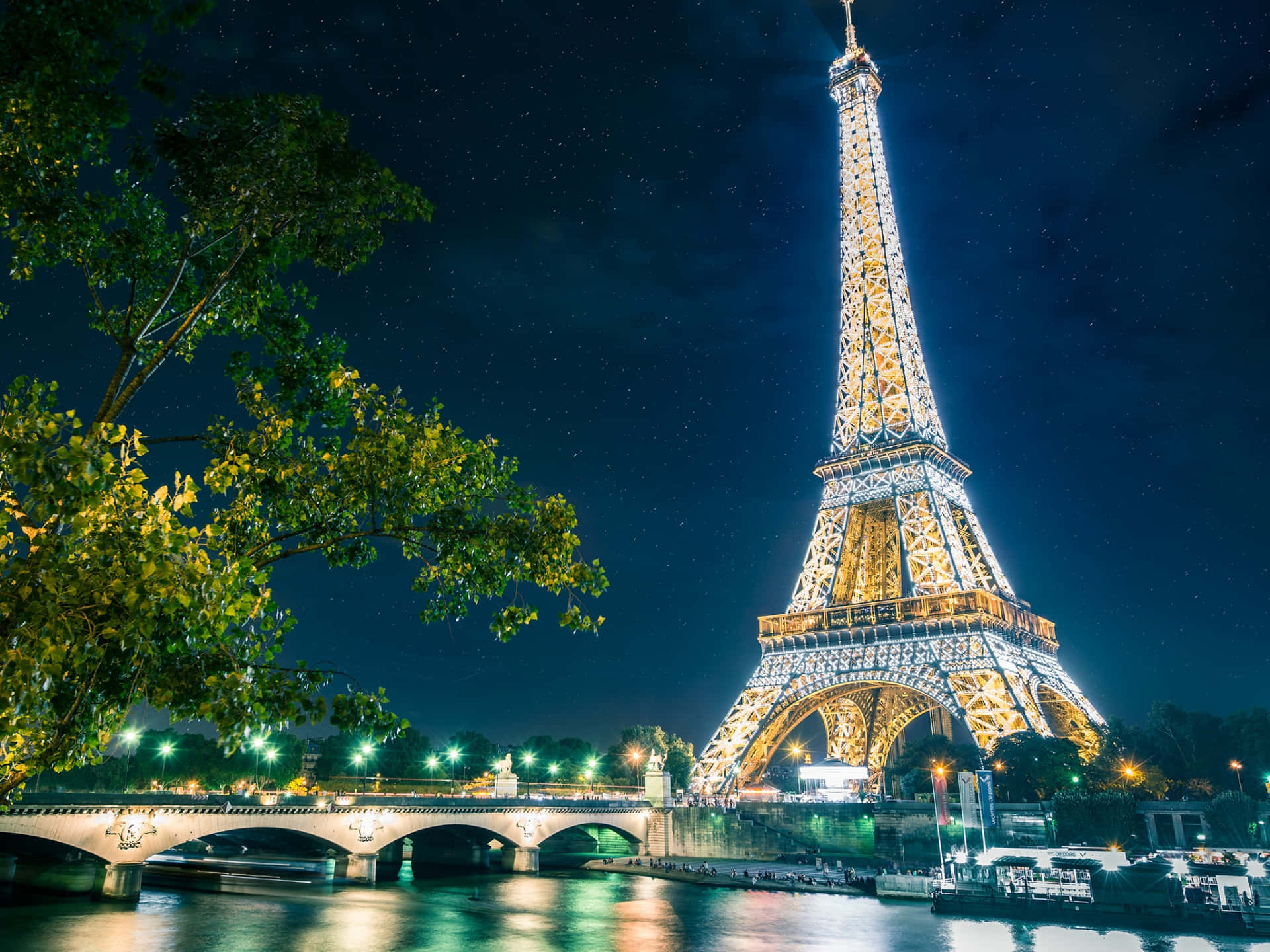 Eiffel Tower 桌布 壁紙 — 艾菲爾鐵塔在繁星點點的夜空下閃耀，倒映在塞納河中，橋樑和茂密的樹木襯托著這個標誌性的巴黎地標。