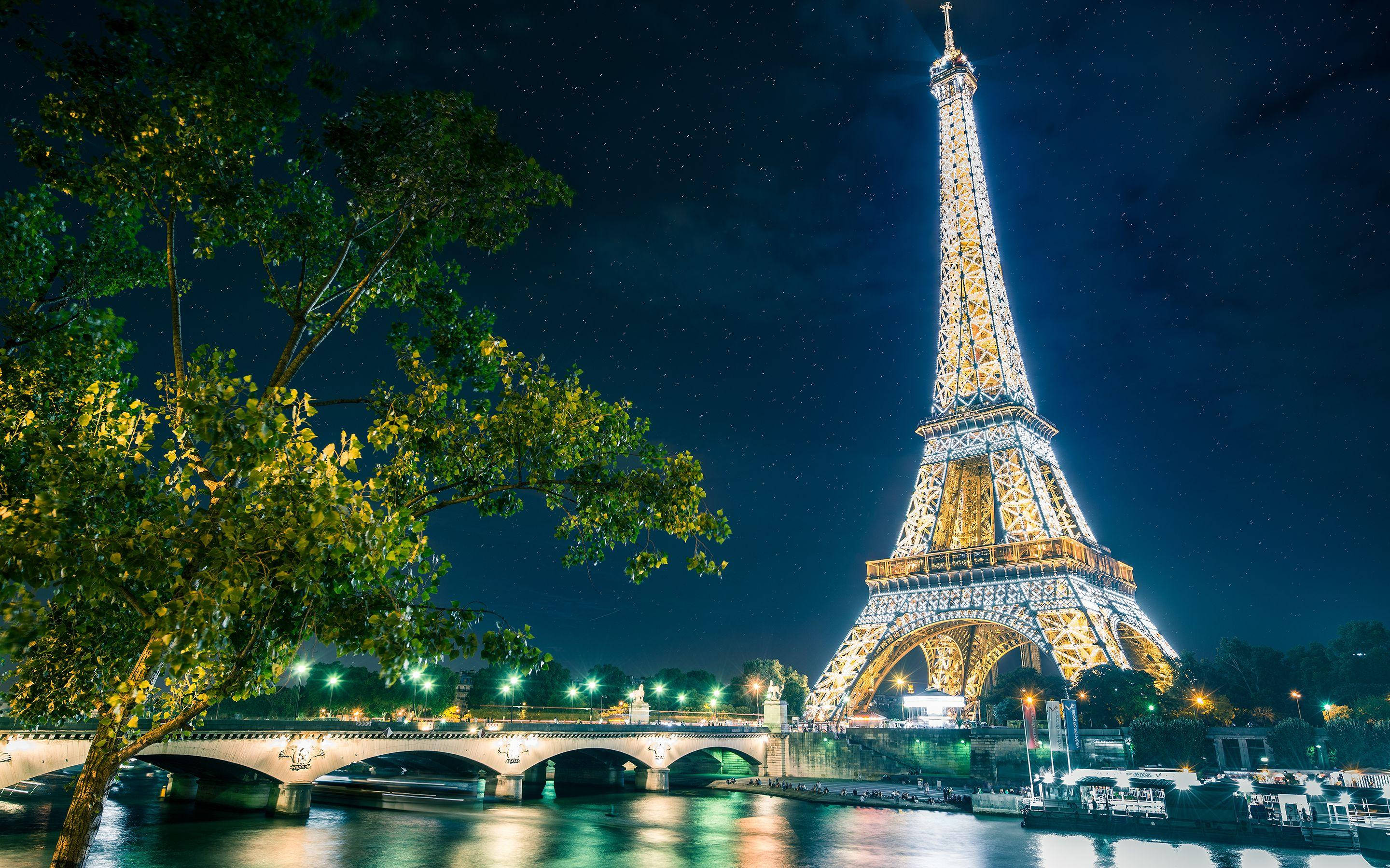 Paris Eiffel Tower 桌布 壁紙 — 艾菲爾鐵塔在繁星點點的夜空下閃閃發光，倒映在塞納河中，前景是一座綠樹成蔭的橋樑。