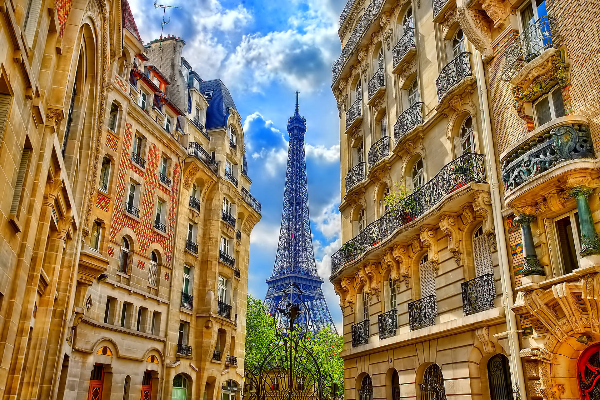 Eiffel Tower 桌布 壁紙 — 艾菲爾鐵塔在裝飾華麗的巴黎建築之間升起，背景是明亮的藍天和蓬鬆的白雲。