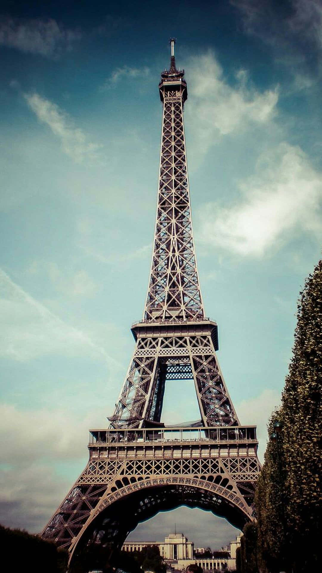 Paris Aesthetic 桌布 壁紙 — 艾菲爾鐵塔在部分多雲的天空下高高聳立，茂密的綠樹環繞其底部。