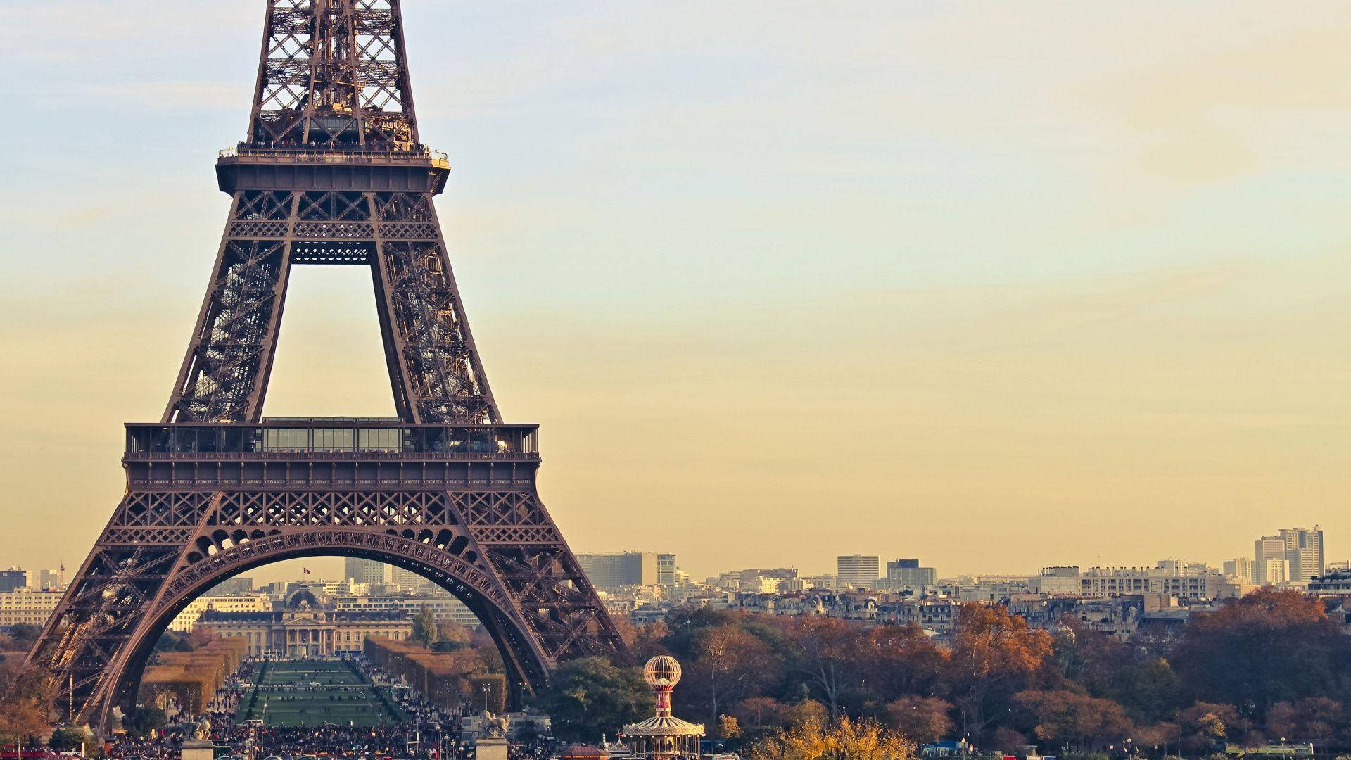 Eiffel Tower 桌布 壁紙 — 艾菲爾鐵塔在金色夕陽天空下高聳矗立，下方是秋天的樹木和巴黎的屋頂。
