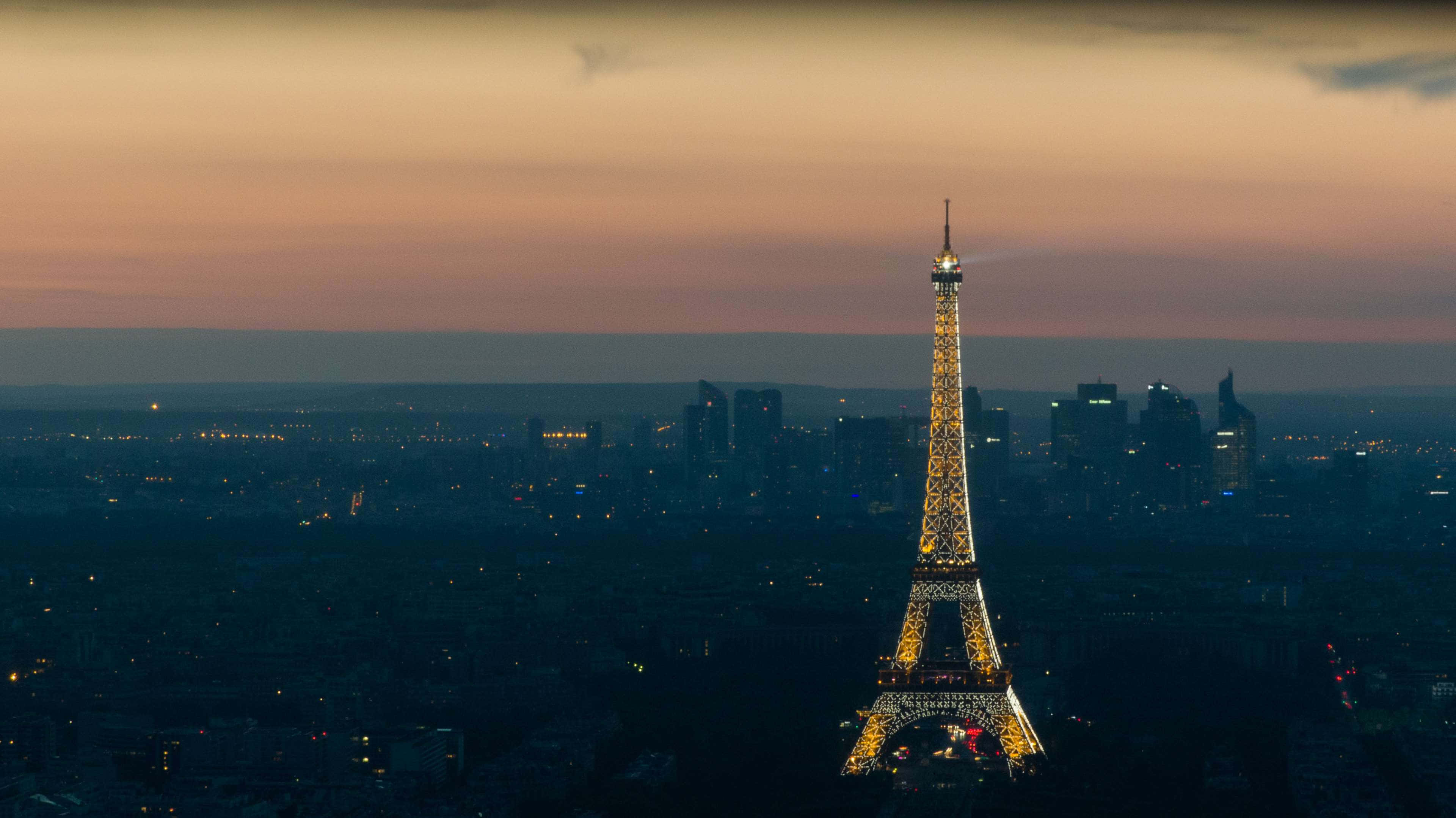 Paris At Night 桌布 壁紙 — 艾菲爾鐵塔在黃昏的粉紅和橙色天空下閃耀著金色光芒，巴黎的城市燈光在下方閃爍。