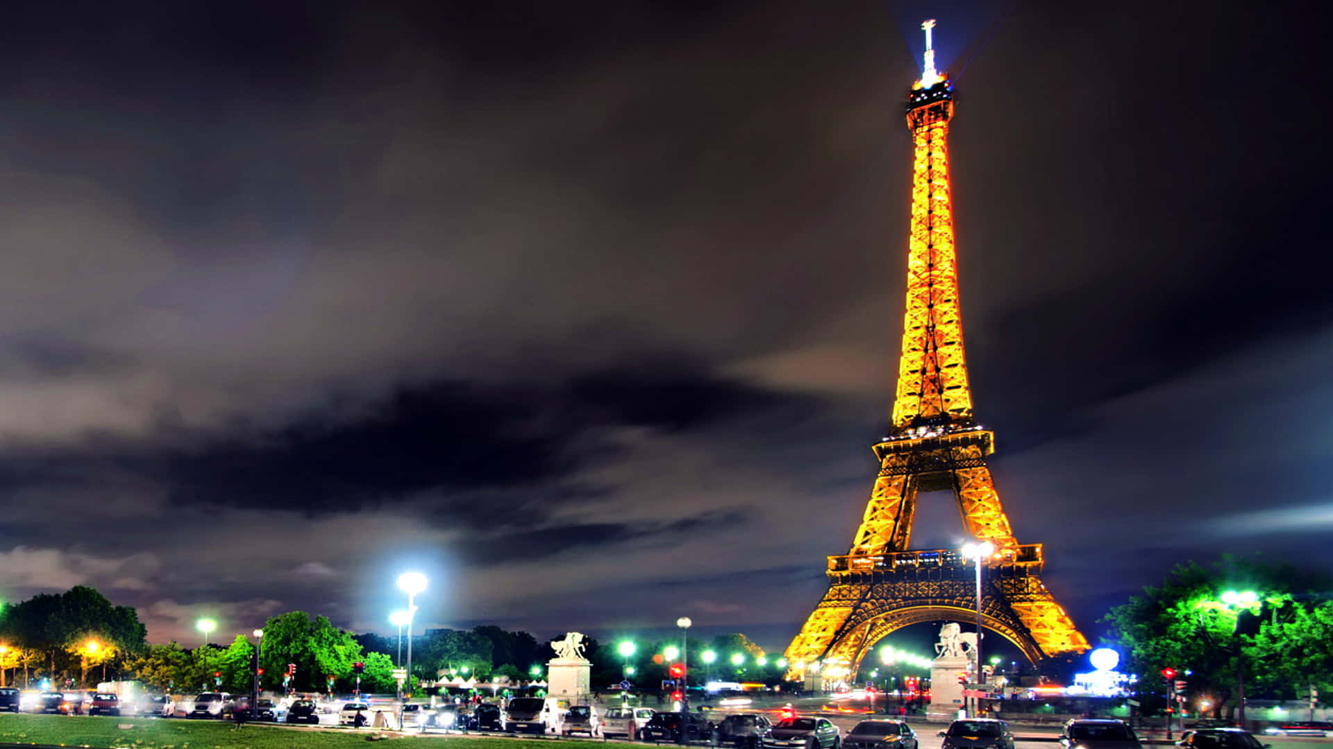 Eiffel Tower 桌布 壁紙 — 艾菲爾鐵塔在黑暗多雲的夜空下溫暖地發光，下方城市燈光和交通模糊不清。