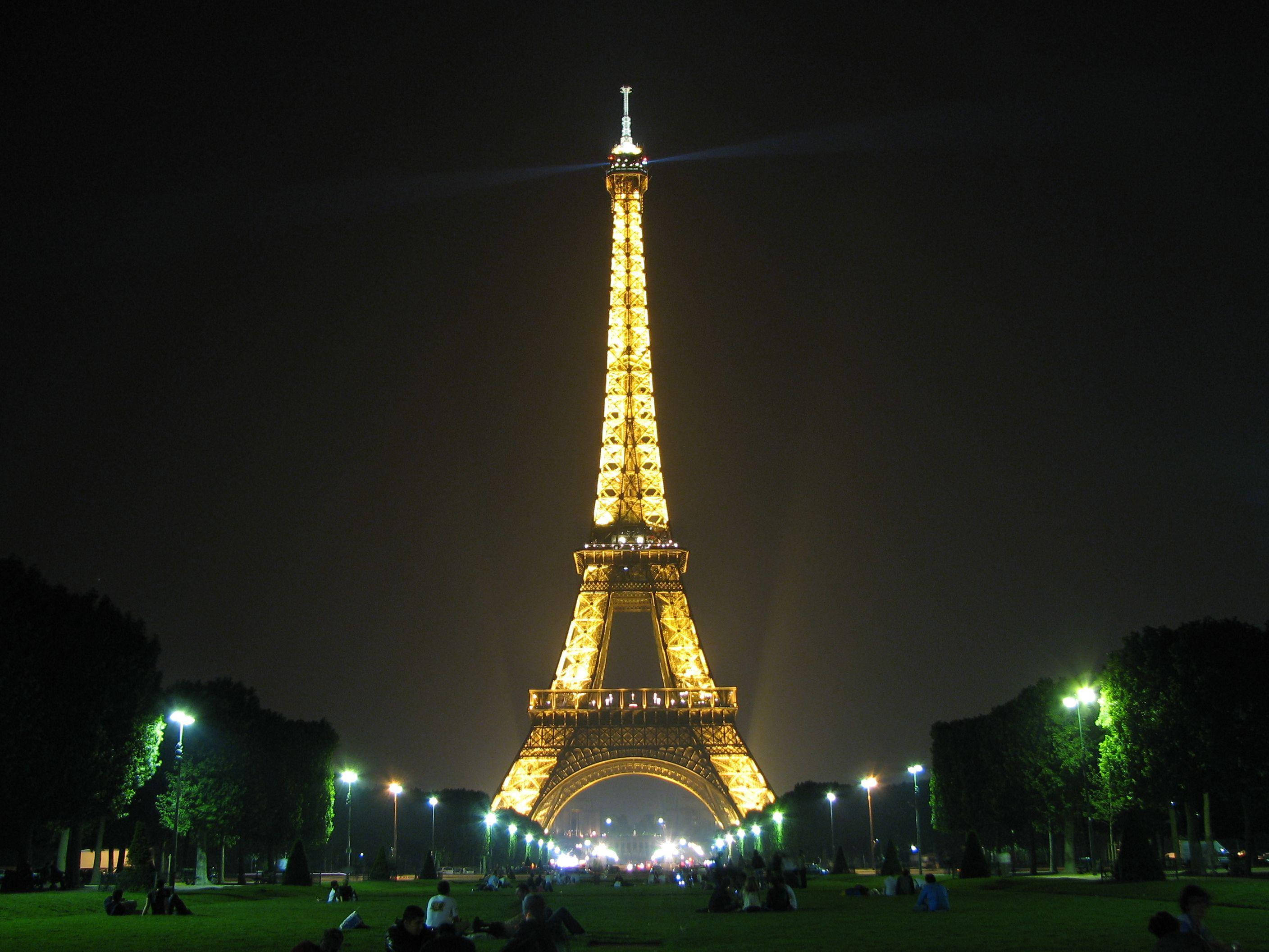 Eiffel Tower 桌布 壁紙 — 艾菲爾鐵塔在黑暗的夜空中閃耀著金色光芒，人們在下面的草地上放鬆。