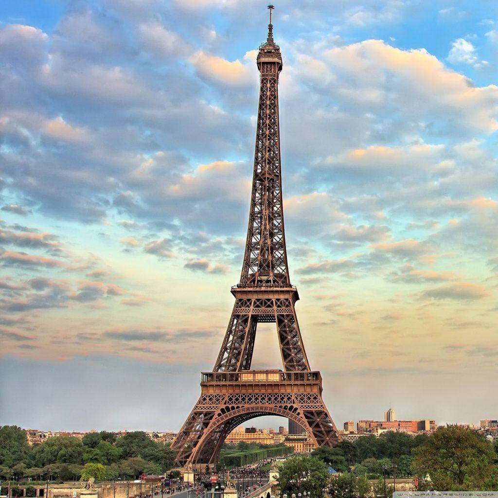 Eiffel Tower 桌布 壁紙 — 艾菲爾鐵塔高聳入雲，背景是柔和的金色天空，散落著雲朵和遠處的巴黎天際線。