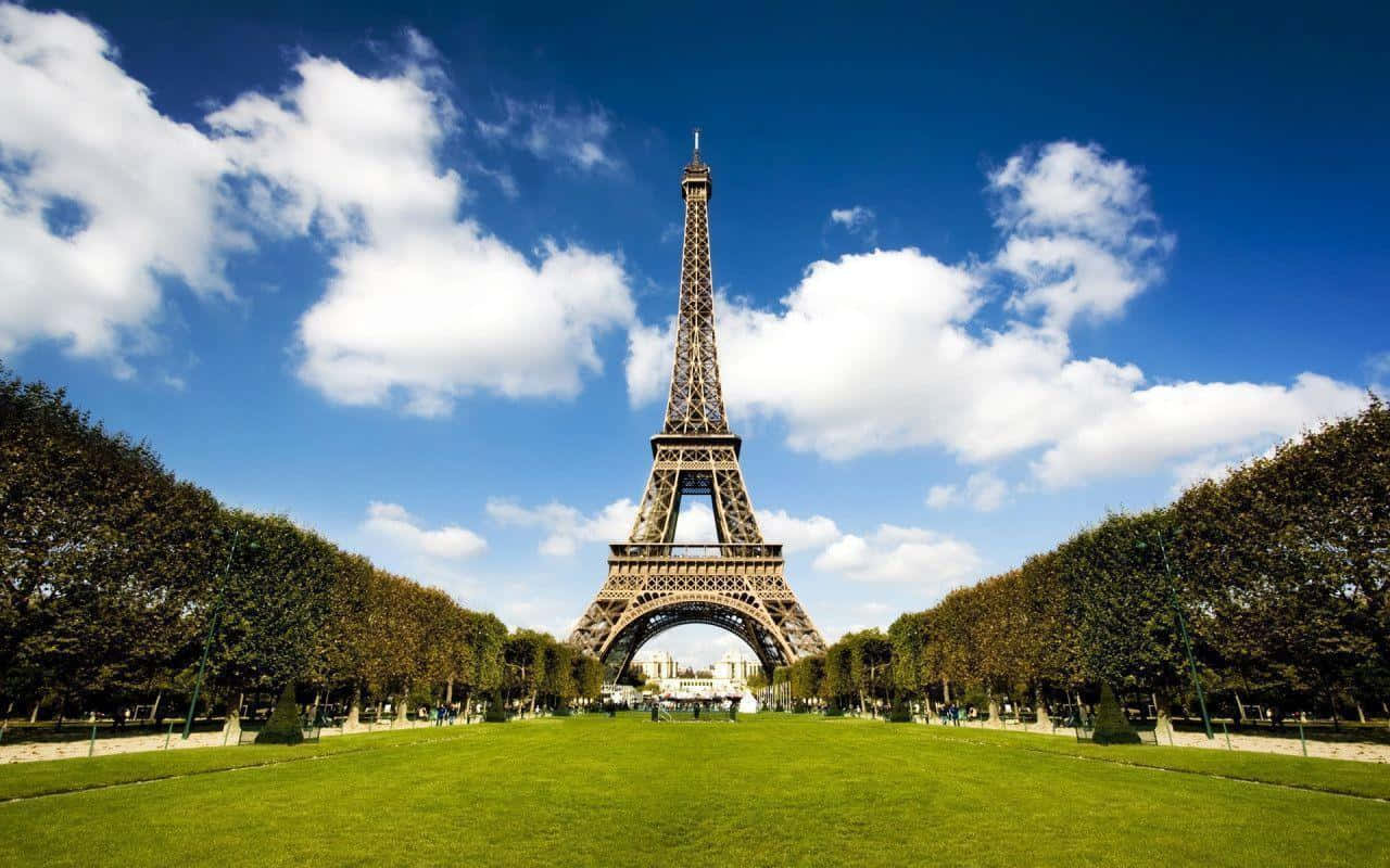Eiffel Tower 桌布 壁紙 — 艾菲爾鐵塔高聳入雲，襯著點綴著蓬鬆白雲的鮮豔藍天，兩旁對稱排列著茂密的綠樹和修剪整齊的草坪。