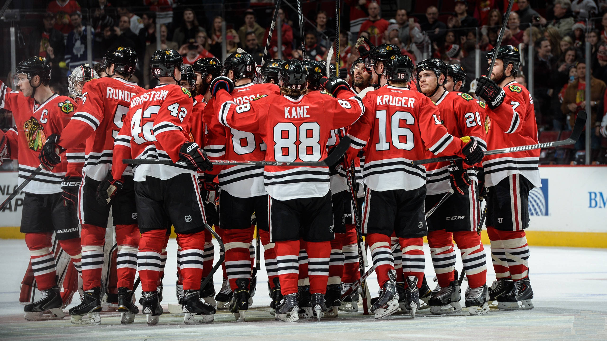 Blackhawks 桌布 壁紙 — 芝加哥黑鷹隊的冰球運動員身穿紅色球衣，在比賽中緊密聚集在冰上，背景有球迷。