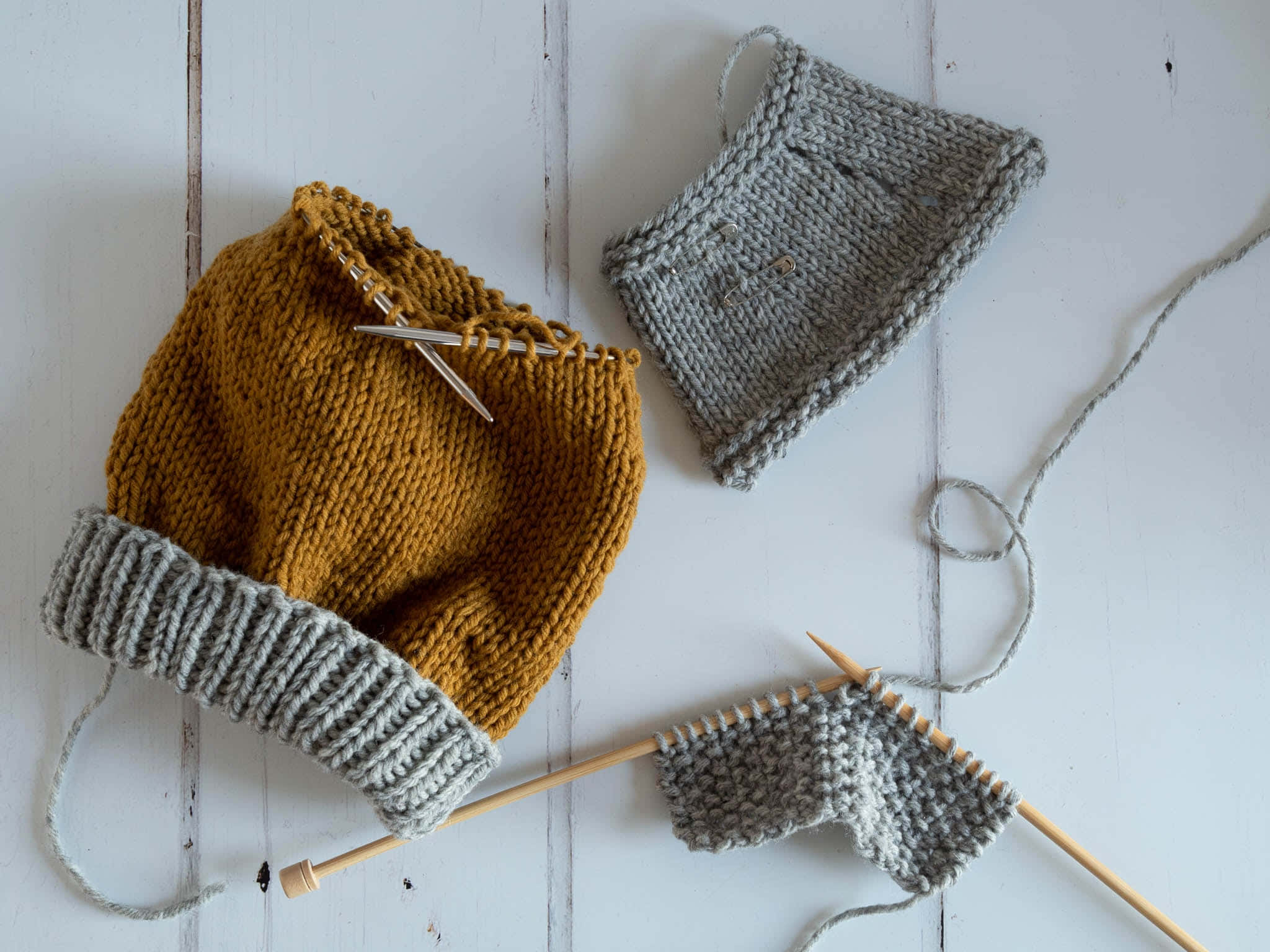 Knitting 桌布 壁紙 — 芥末黃色和灰色的針織帽，針和毛線散落在白色木質表面上。
