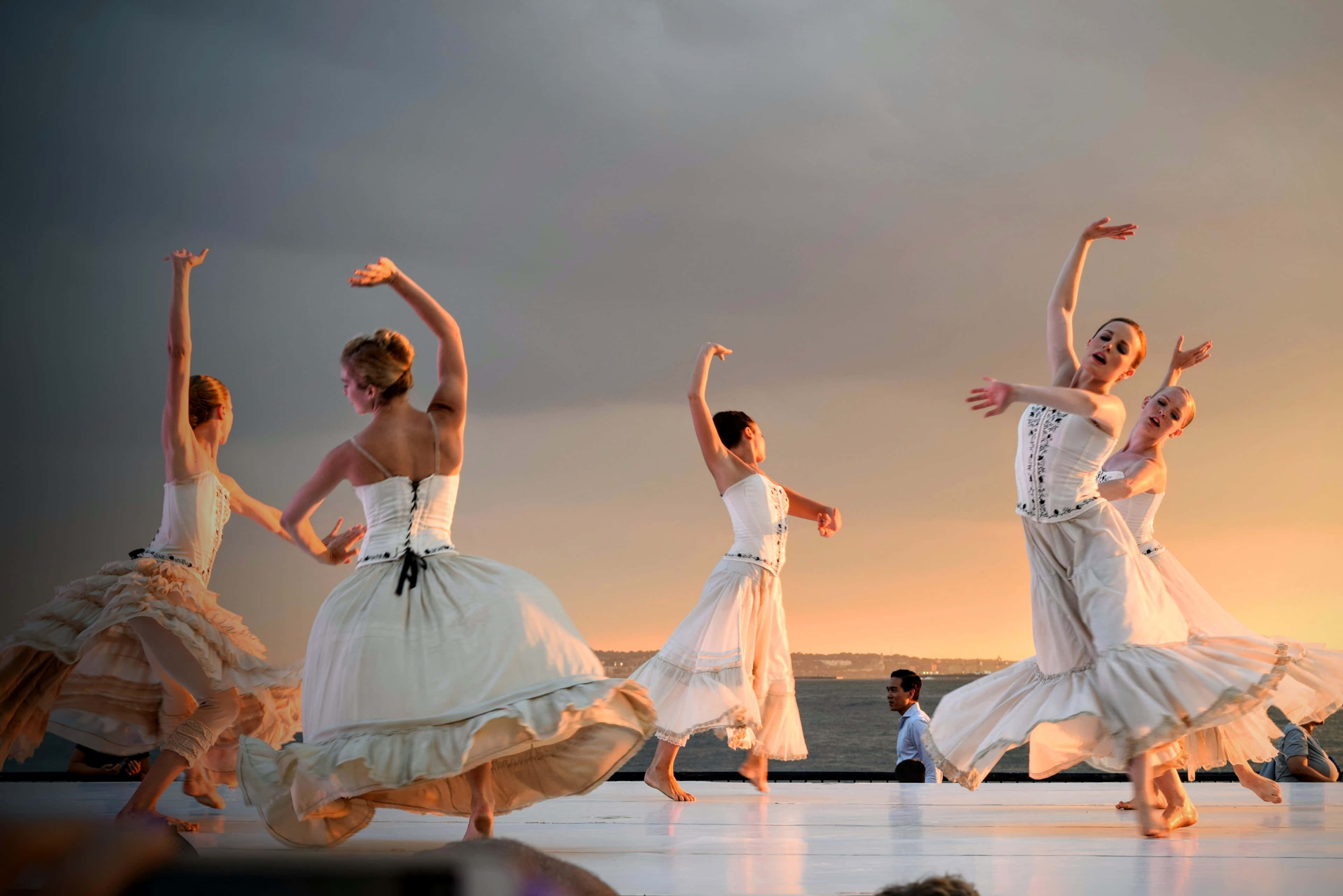 Ballet 桌布 壁紙 — 芭蕾舞者身穿白色服裝，在日落時分的舞台上優雅表演，背景是城市天際線。