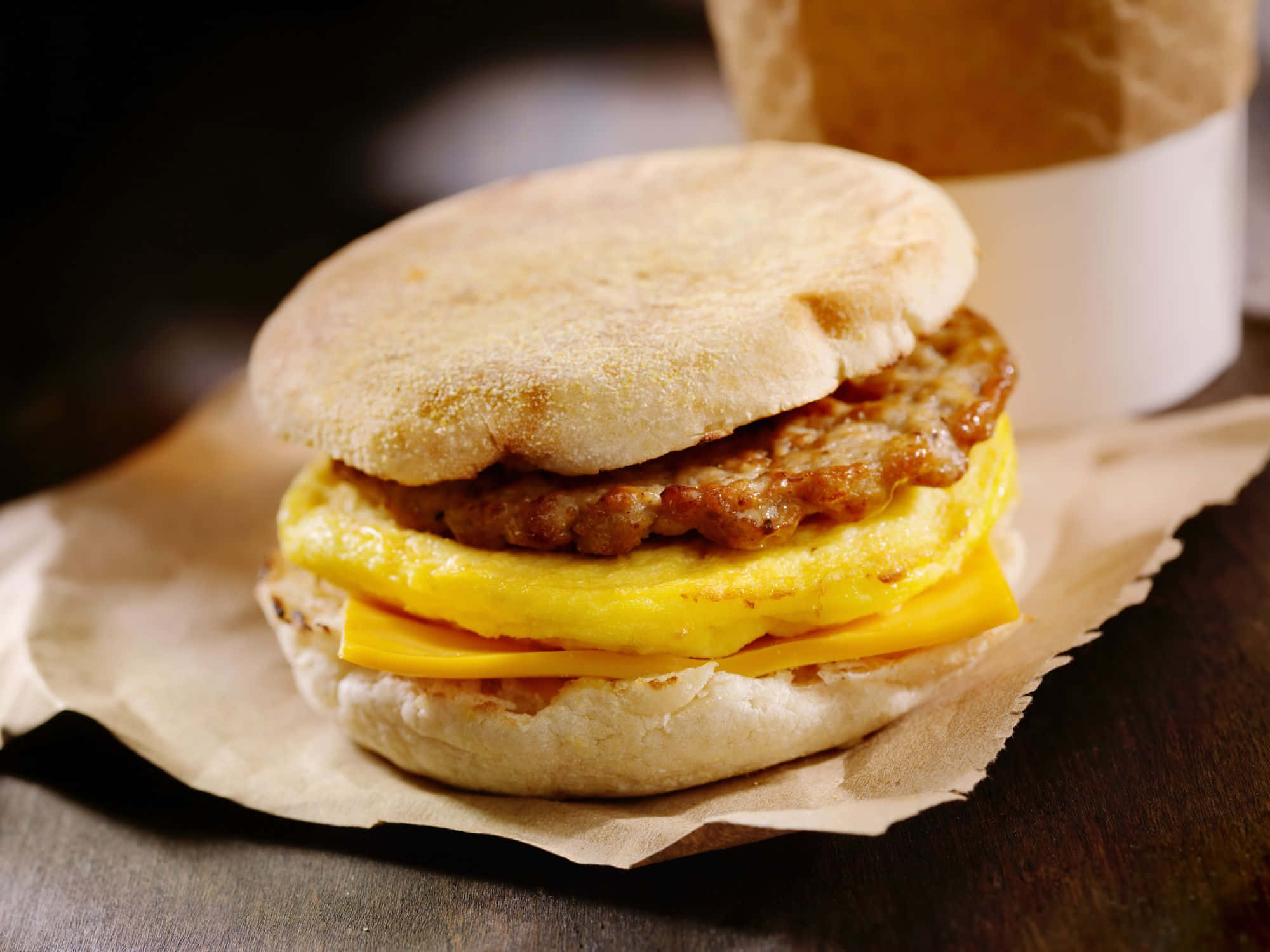 fast food 桌布 壁紙 — 英式鬆餅早餐三明治的特寫，內有香腸肉餅、炒蛋和起司，放在烘焙紙上。