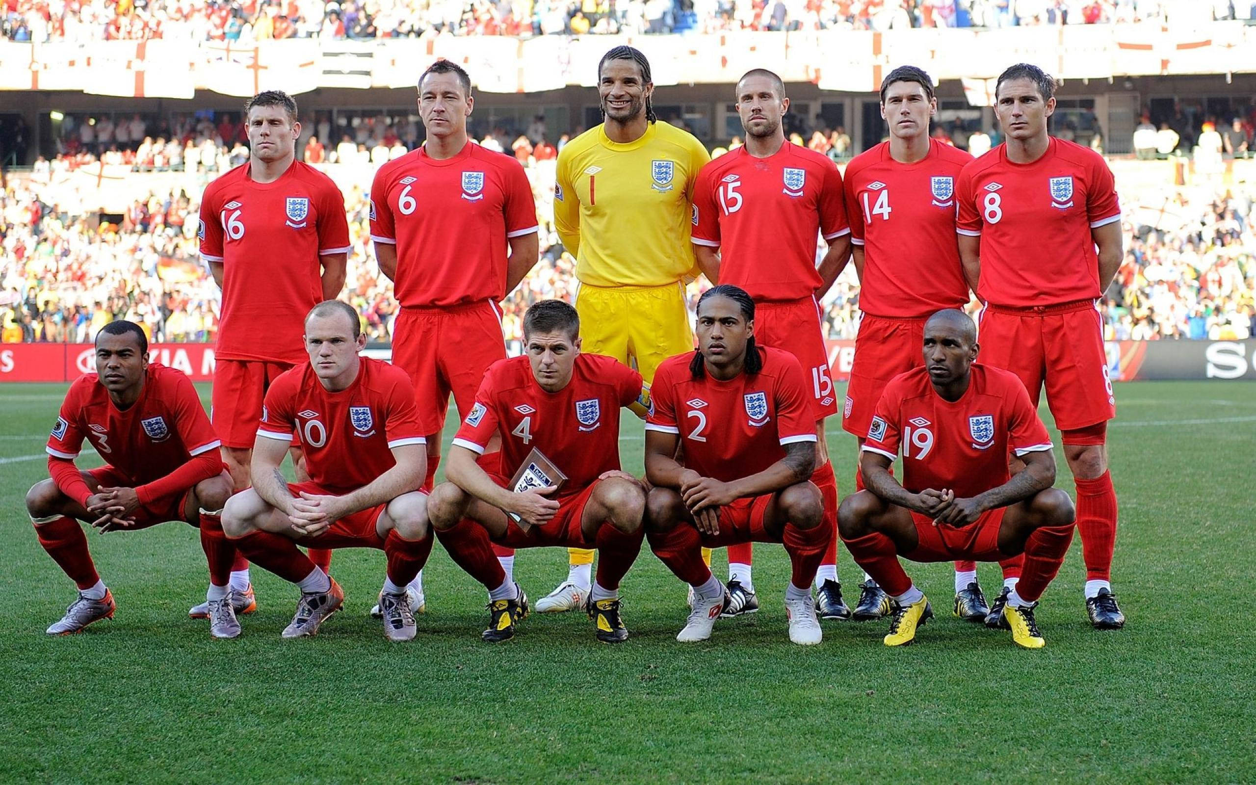 Football Players 桌布 壁紙 — 英格蘭國家足球隊身穿紅色球衣，在陽光明媚的綠色球場上擺姿勢，背景是體育場觀眾。