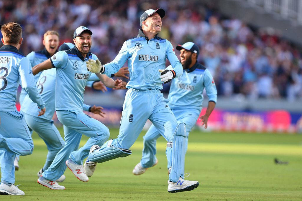 England Cricket 桌布 壁紙 — 英格蘭板球球員身穿淺藍色球衣，在綠色球場上興奮慶祝，背景是模糊的人群。