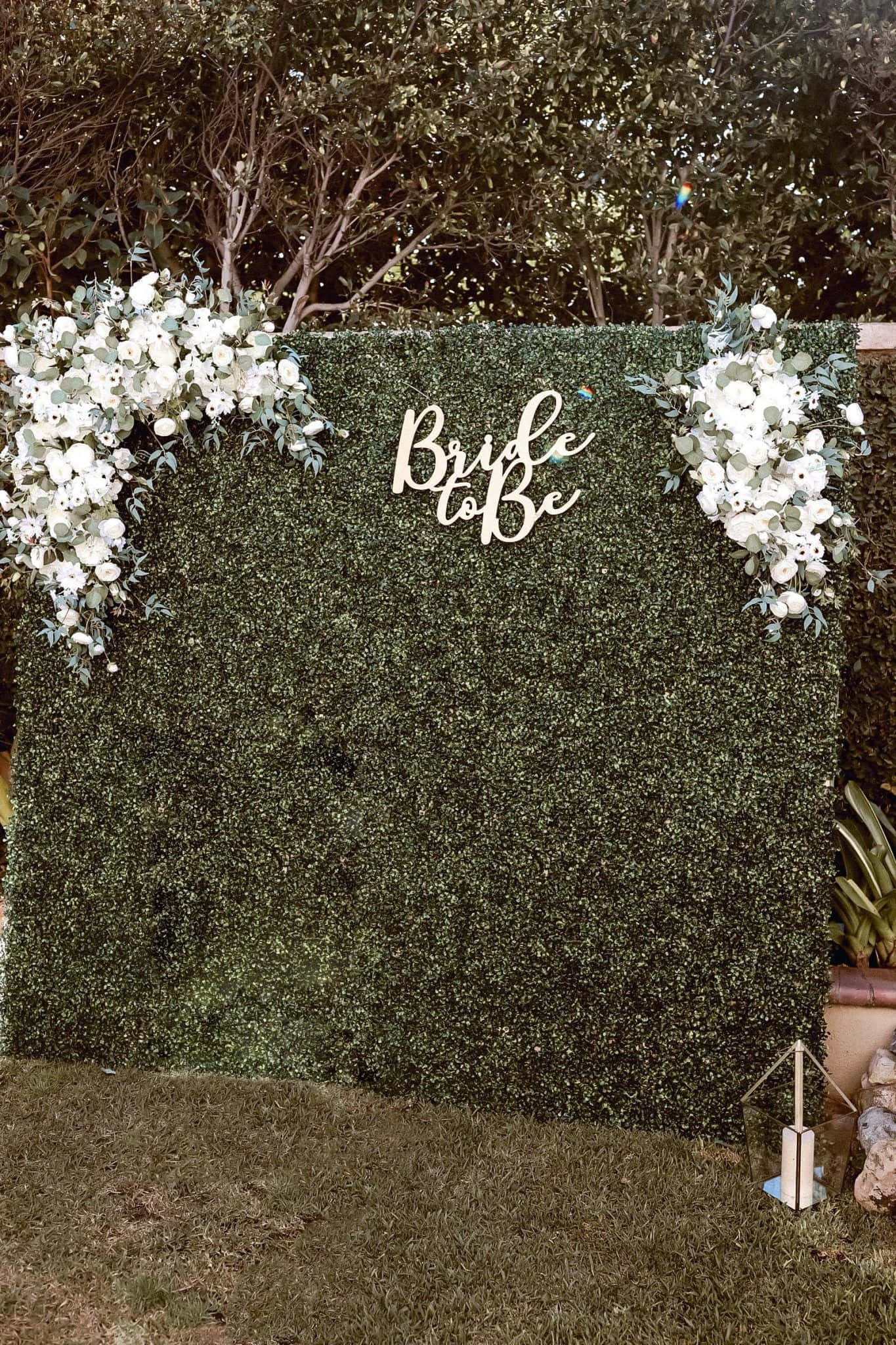 Bride To Be 桌布 壁紙 — 茂密的綠色樹籬牆，飾有白色花朵和「Bride to Be」標誌，用作花園婚禮背景。
