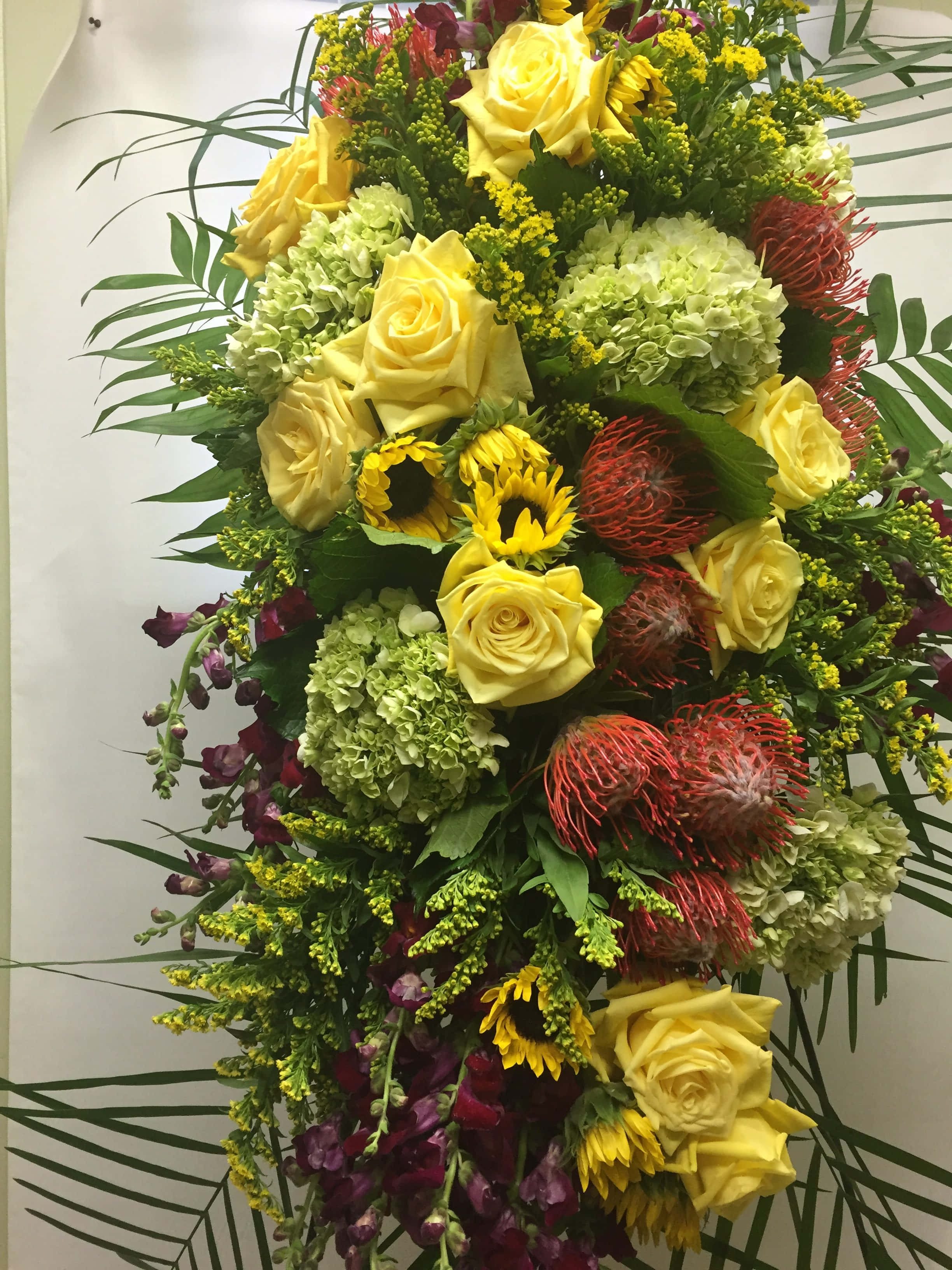 Funeral Flower Arrangements 桌布 壁紙 — 茂盛的花卉佈置，以黃玫瑰、向日葵、綠色繡球花和紅色帝王花層疊而下，並以棕櫚葉框住。