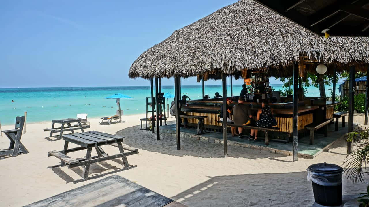 Jamaican Beach 桌布 壁紙 — 茅草屋頂海灘酒吧，白色沙灘上擺放木製野餐桌，旁邊是碧綠海洋，上方是清澈藍天。