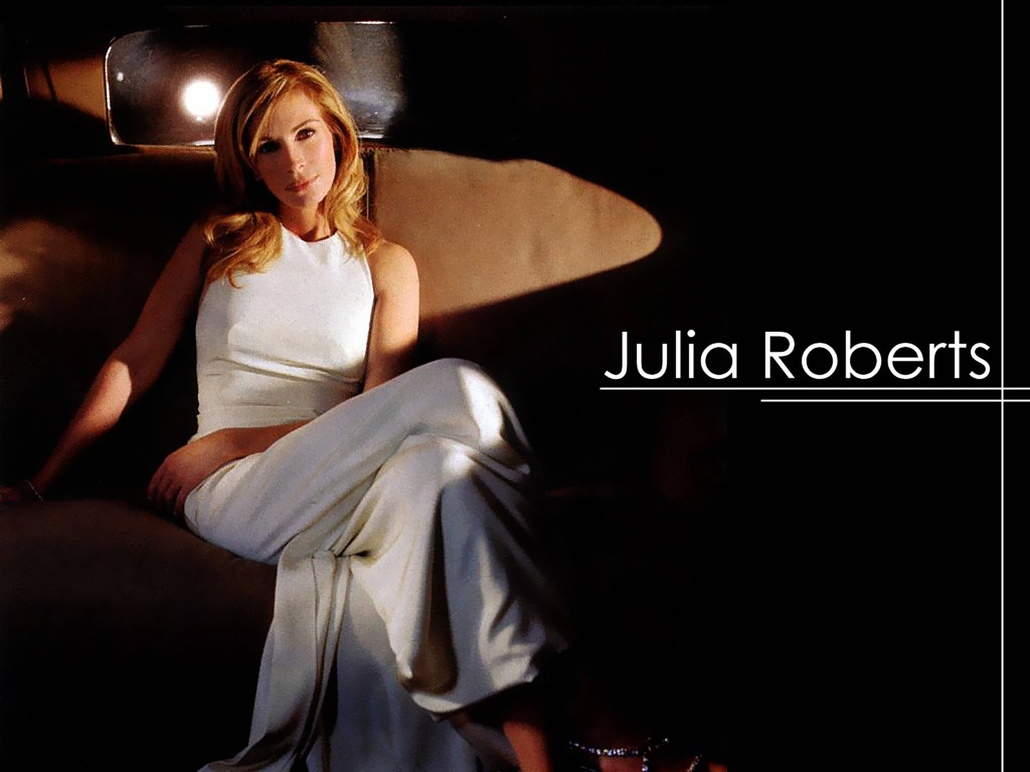 Julia Roberts 桌布 壁紙 — 茱莉亞羅勃茲身穿白色連衣裙，在溫暖的聚光燈下優雅地坐著，背景是昏暗的室內。