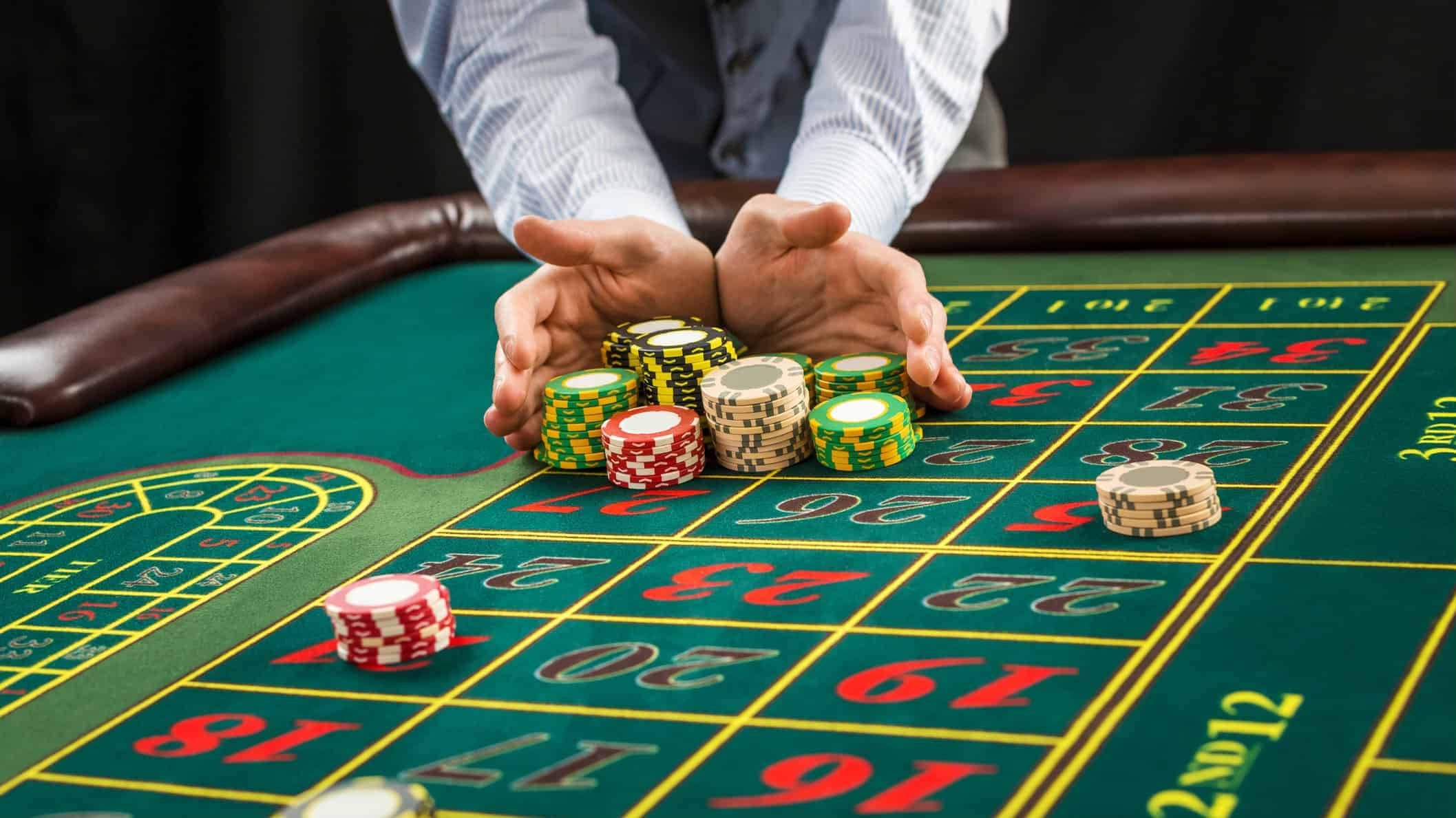 Poker 桌布 壁紙 — 荷官的手在綠色輪盤桌上的彩色賭場籌碼上方盤旋，桌上帶有編號的投注格。