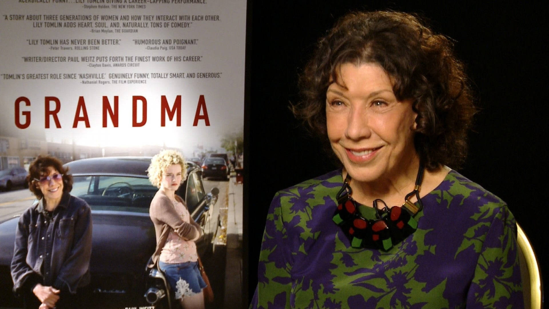 女演員 桌布 壁紙 — 莉莉湯姆林在電影《Grandma》海報前溫暖微笑，海報上有她和一位年輕女子站在車旁。