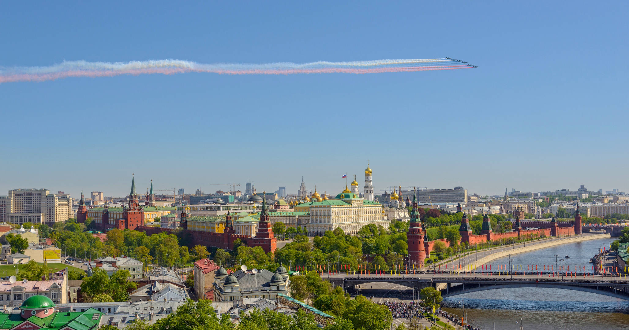 Russia 桌布 壁紙 — 莫斯科克里姆林宮的鳥瞰圖，有紅磚牆、金色圓頂和一座橫跨河流的橋樑，噴射機在上空飛過留下彩色煙霧痕跡。