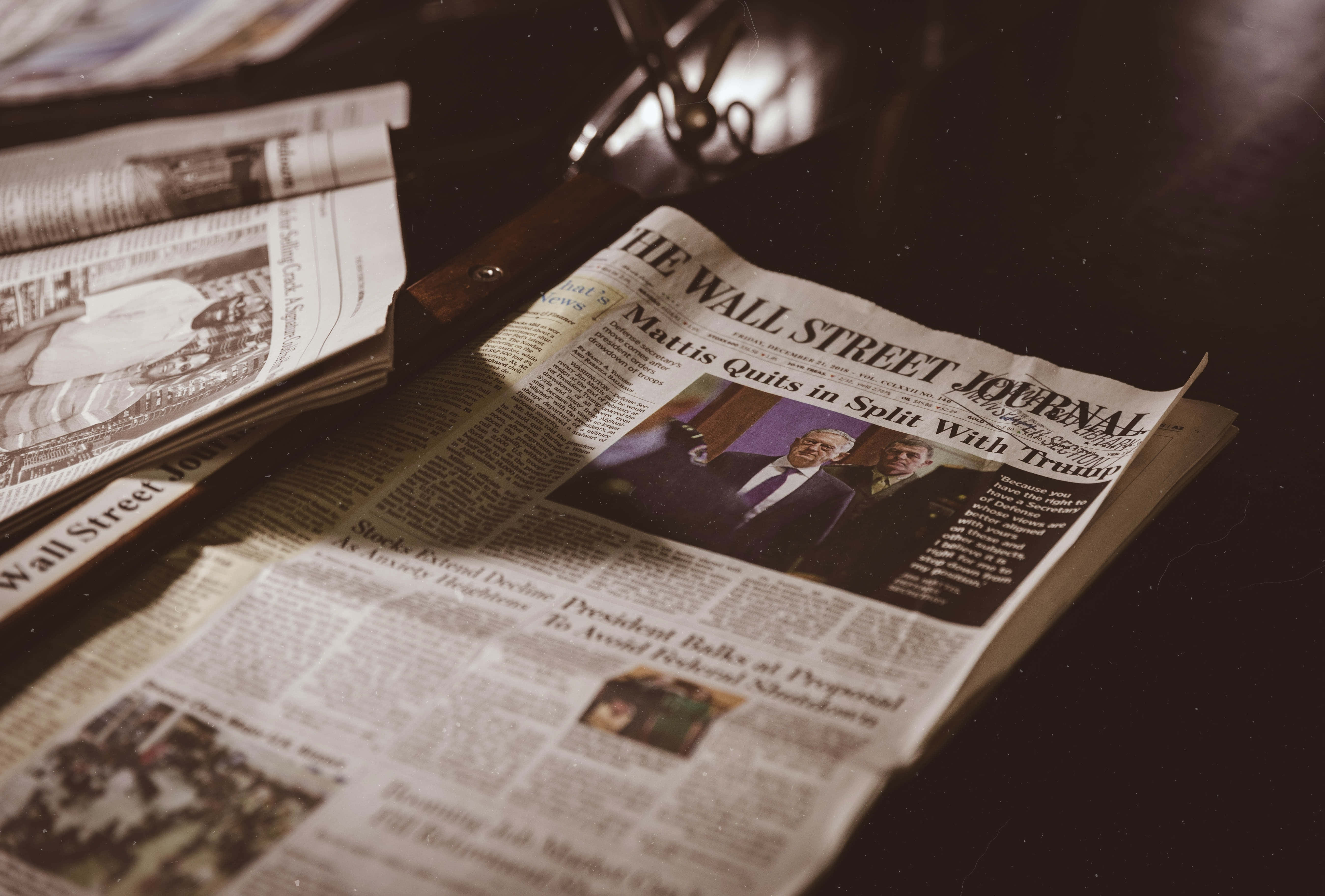Old Newspaper 桌布 壁紙 — 《華爾街日報》報紙的特寫，帶有標題和照片，放在溫暖光線下的深色木質表面上。