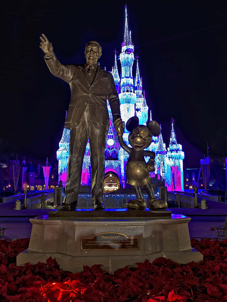 Disneyland Castle 桌布 壁紙 — 華特迪士尼和米奇老鼠的銅像在夜晚於一座發光的藍紫色城堡旁揮手，周圍環繞著紅色聖誕紅。