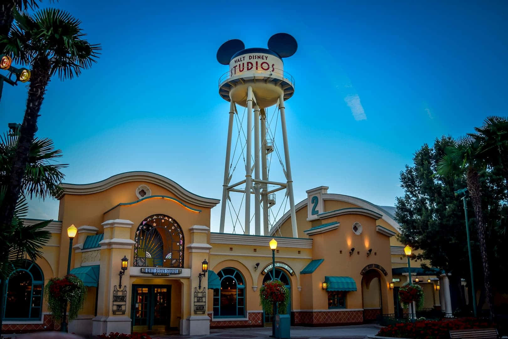Walt Disney Studios Motion 桌布 壁紙 — 華特迪士尼工作室的水塔，帶有米奇老鼠耳朵，在暮色天空下矗立於主題建築和棕櫚樹之上。