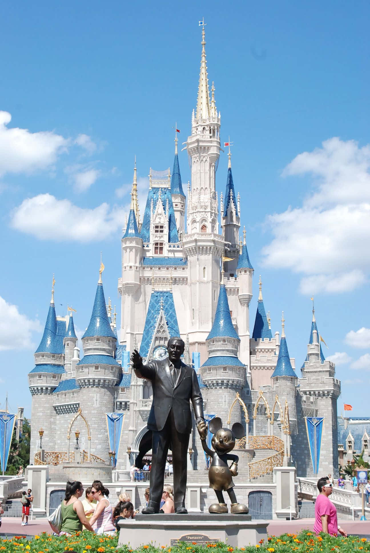 Disney Castle 桌布 壁紙 — 華特迪士尼抱著米奇老鼠的銅像，矗立在標誌性的灰姑娘城堡前，背景是晴朗的藍天和蓬鬆的白雲。