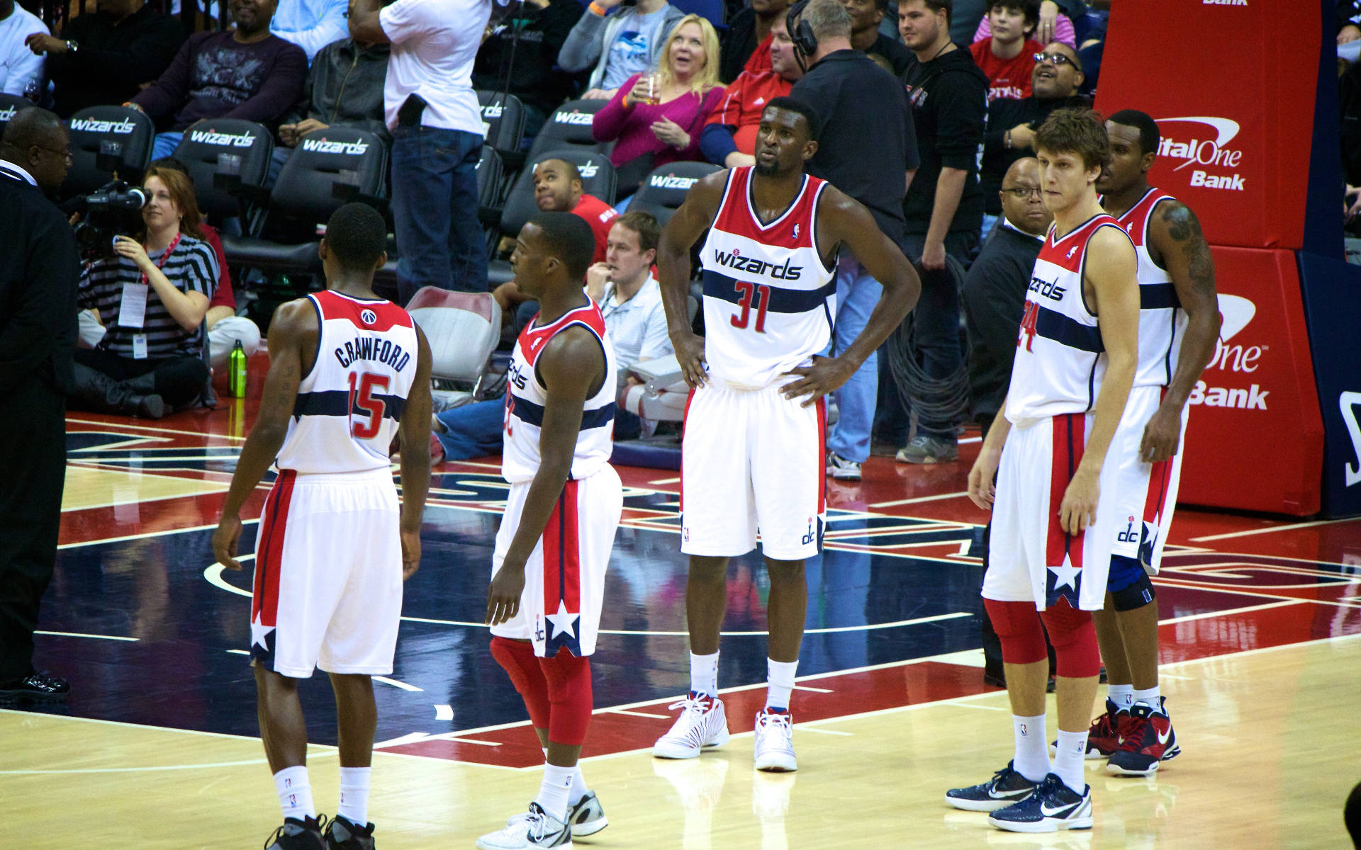 Wizards 桌布 壁紙 — 華盛頓巫師隊籃球運動員身穿白色、紅色和藍色球衣，在比賽期間站在拋光木地板上，背景有球迷。
