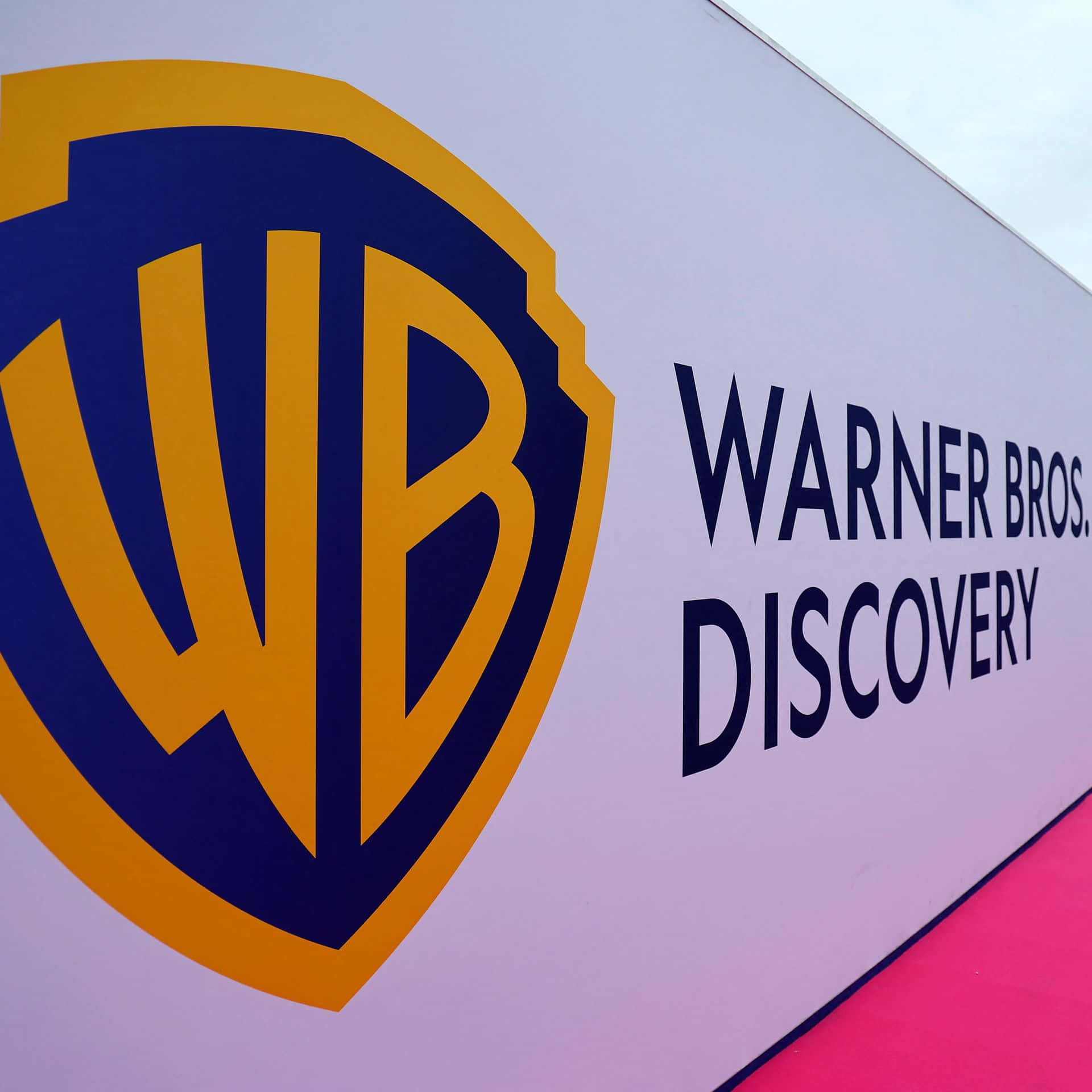 Warner Bros 桌布 壁紙 — 華納兄弟探索公司標誌的特寫，位於白色牆壁上，粉紅色地板，在明亮的日光下。
