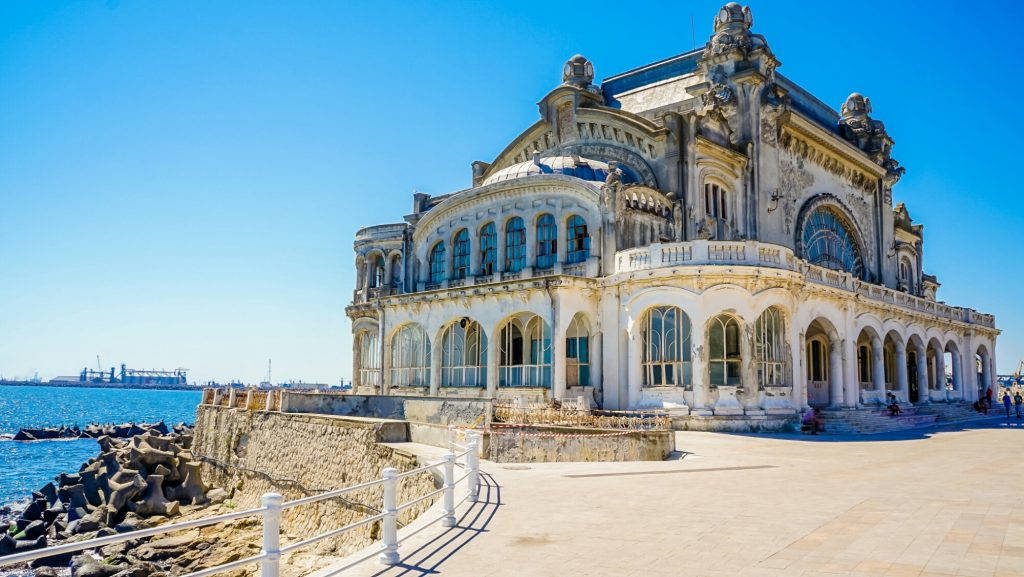 Romania 桌布 壁紙 — 華麗宏偉的海邊劇院，擁有拱形窗戶和圓頂屋頂，在明亮的藍天和寧靜的海洋中顯得飽經風霜。