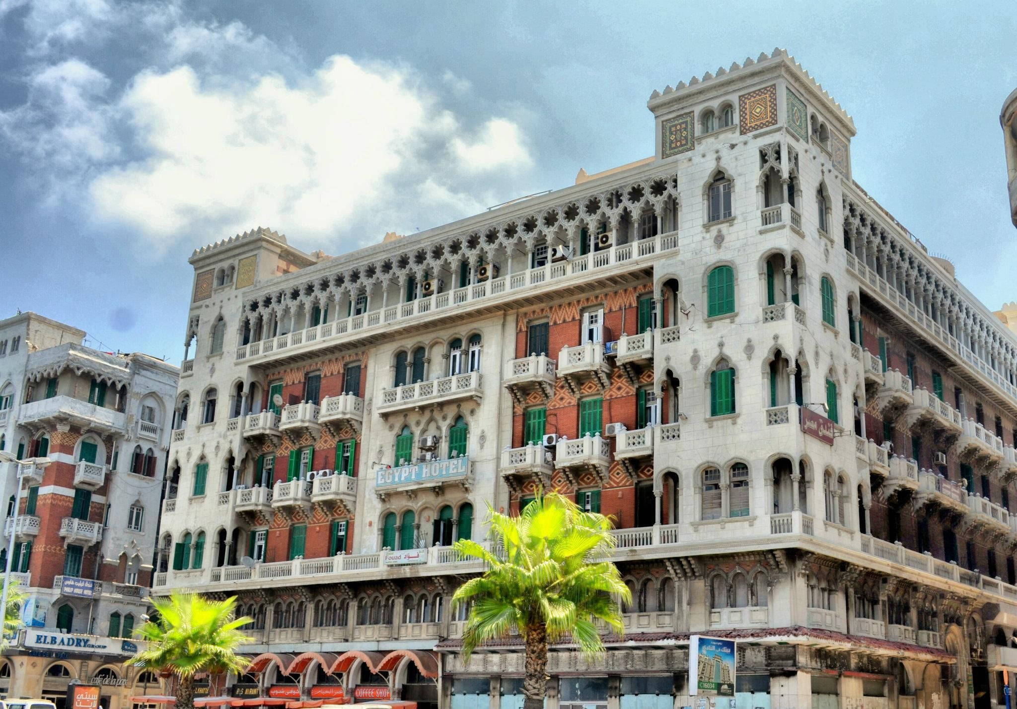 Alexandria 桌布 壁紙 — 華麗的大飯店，配有綠色百葉窗和陽台，矗立在明亮的陰天天空下，前景有棕櫚樹。
