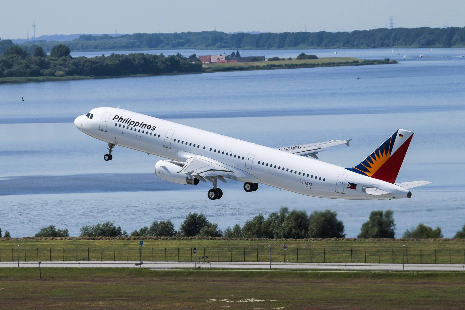 飛機 桌布 壁紙 — 菲律賓航空空中巴士A320在平靜的藍色湖泊上空起飛，周圍有綠樹和遠處的海岸線，天空晴朗。