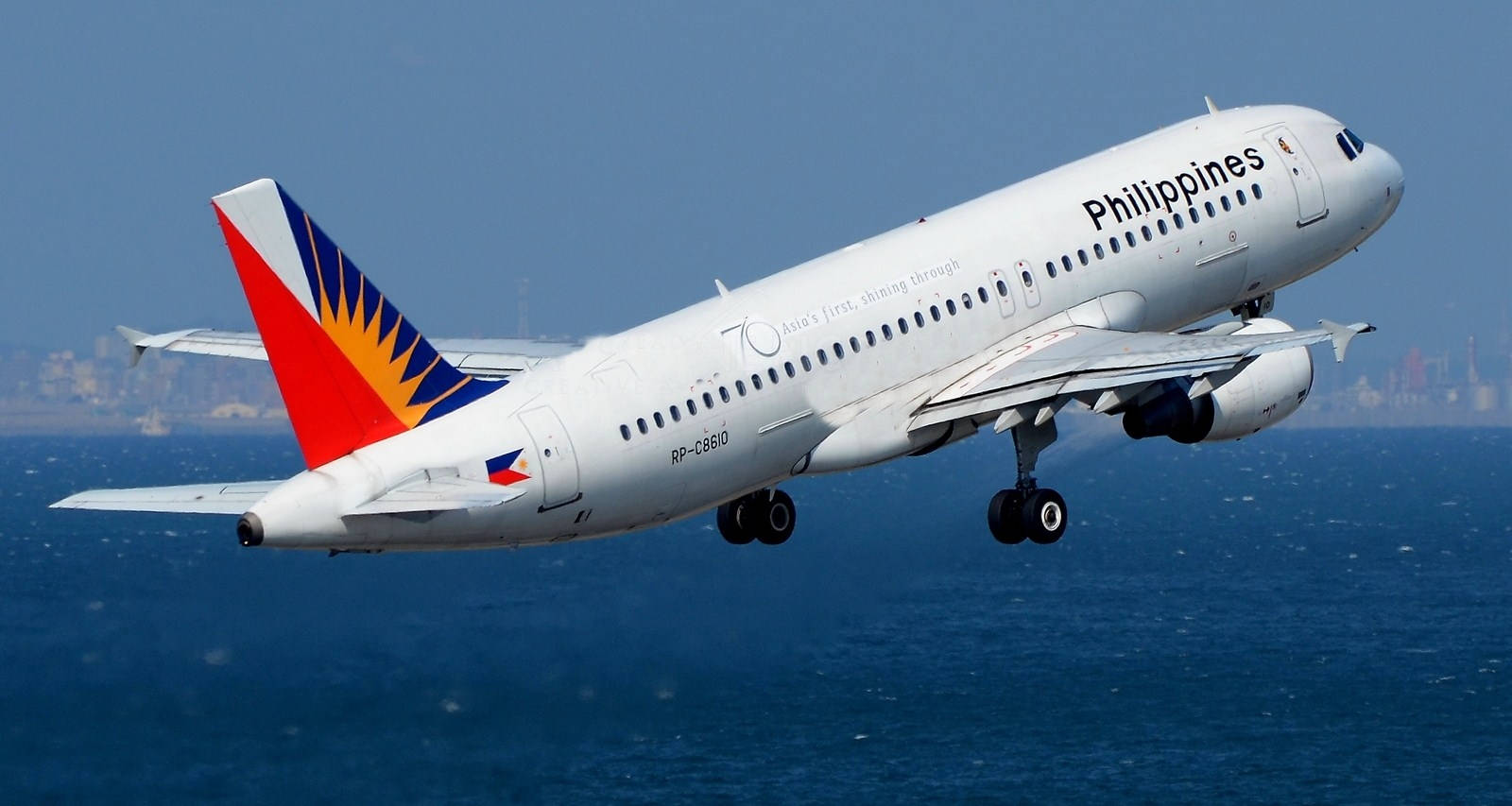 飛機 桌布 壁紙 — 菲律賓航空空中巴士A320放下起落架，在深藍色海洋上空低飛，遠處城市天際線在晴朗天空下。