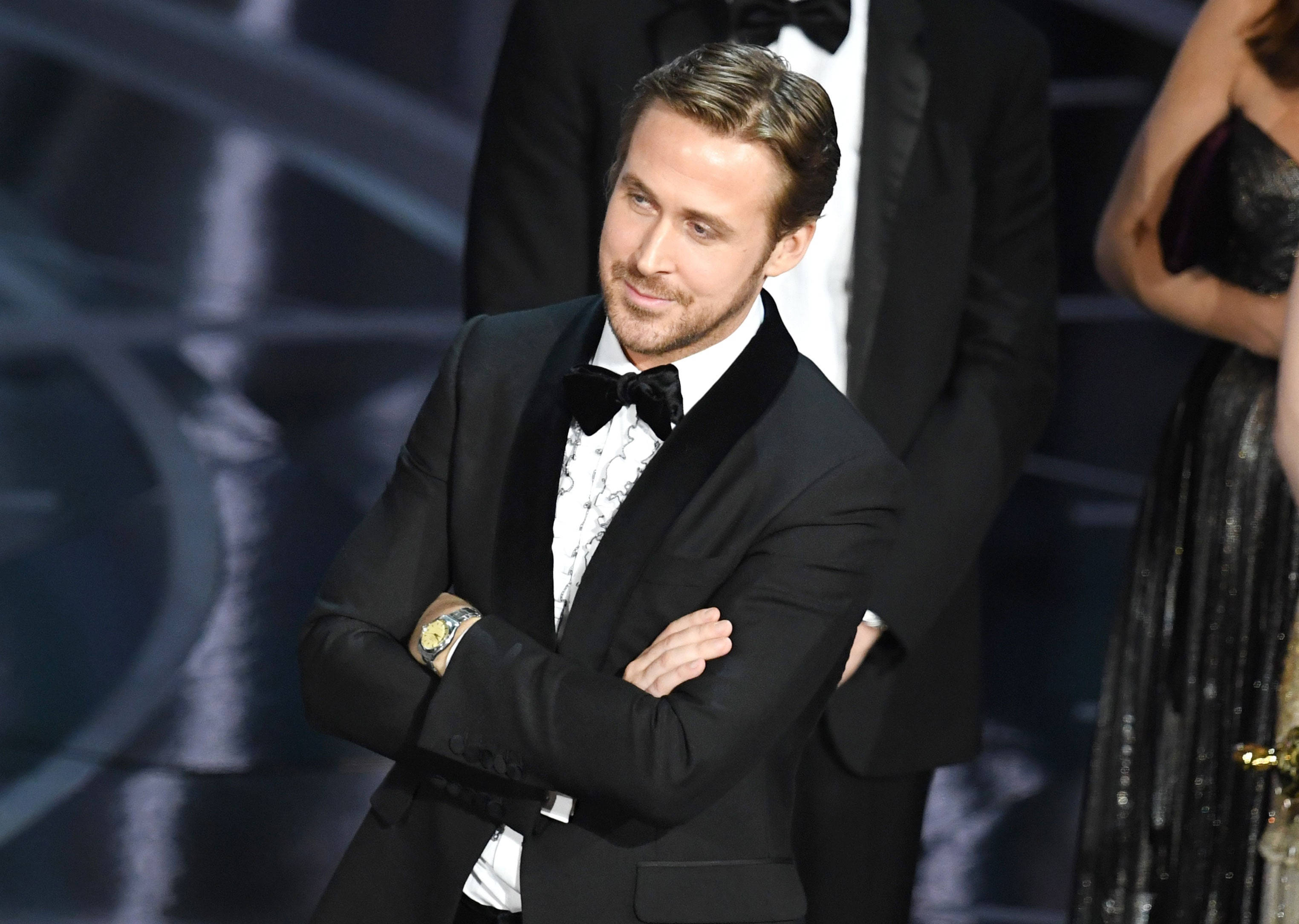 Ryan Gosling 桌布 壁紙 — 萊恩葛斯林自信地穿著黑色燕尾服，雙臂交叉，在明亮的舞台燈光下參加一場華麗的頒獎典禮。