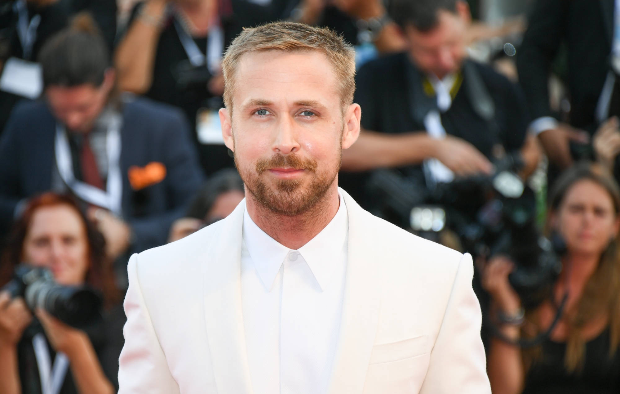 Ryan Gosling 桌布 壁紙 — 萊恩葛斯林身穿筆挺白色西裝，自信地站在紅地毯上，周圍是攝影師和模糊的旁觀者。