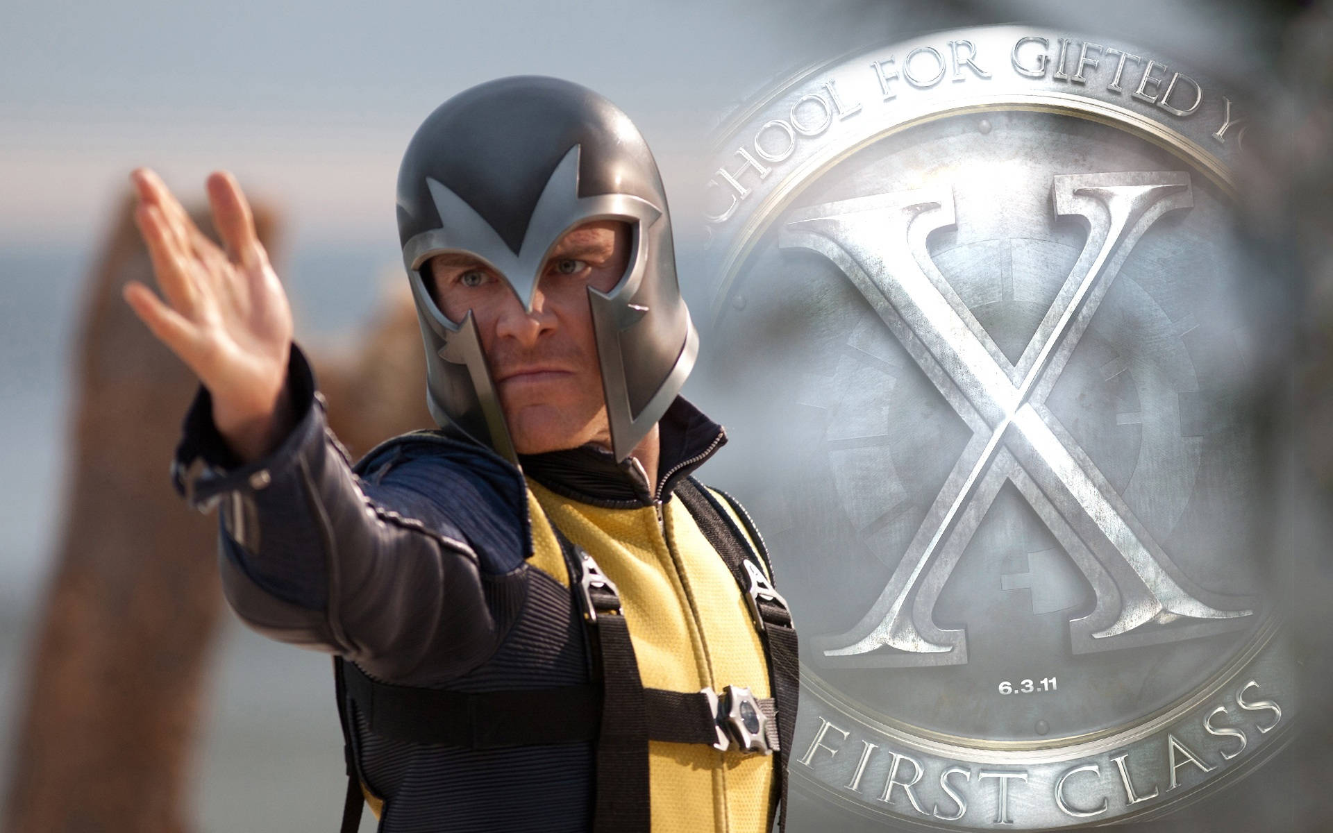 Magneto 桌布 壁紙 — 萬磁王戴著標誌性頭盔，身穿黃黑相間的服裝，在X-Men First Class標誌前伸出手站立。