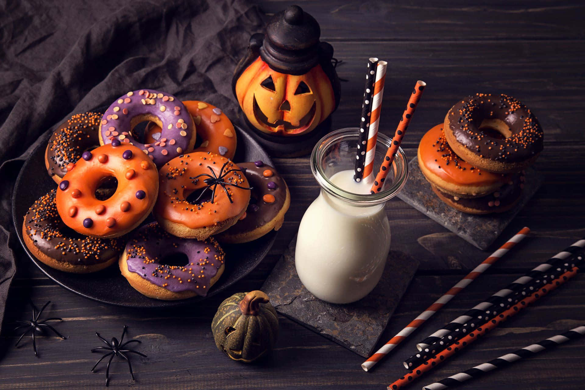 Halloween Treats 桌布 壁紙 — 萬聖節主題的甜甜圈，帶有橙色和紫色糖霜和彩色糖粒，擺放在深色木桌上，配有一個傑克南瓜燈馬克杯和牛奶。