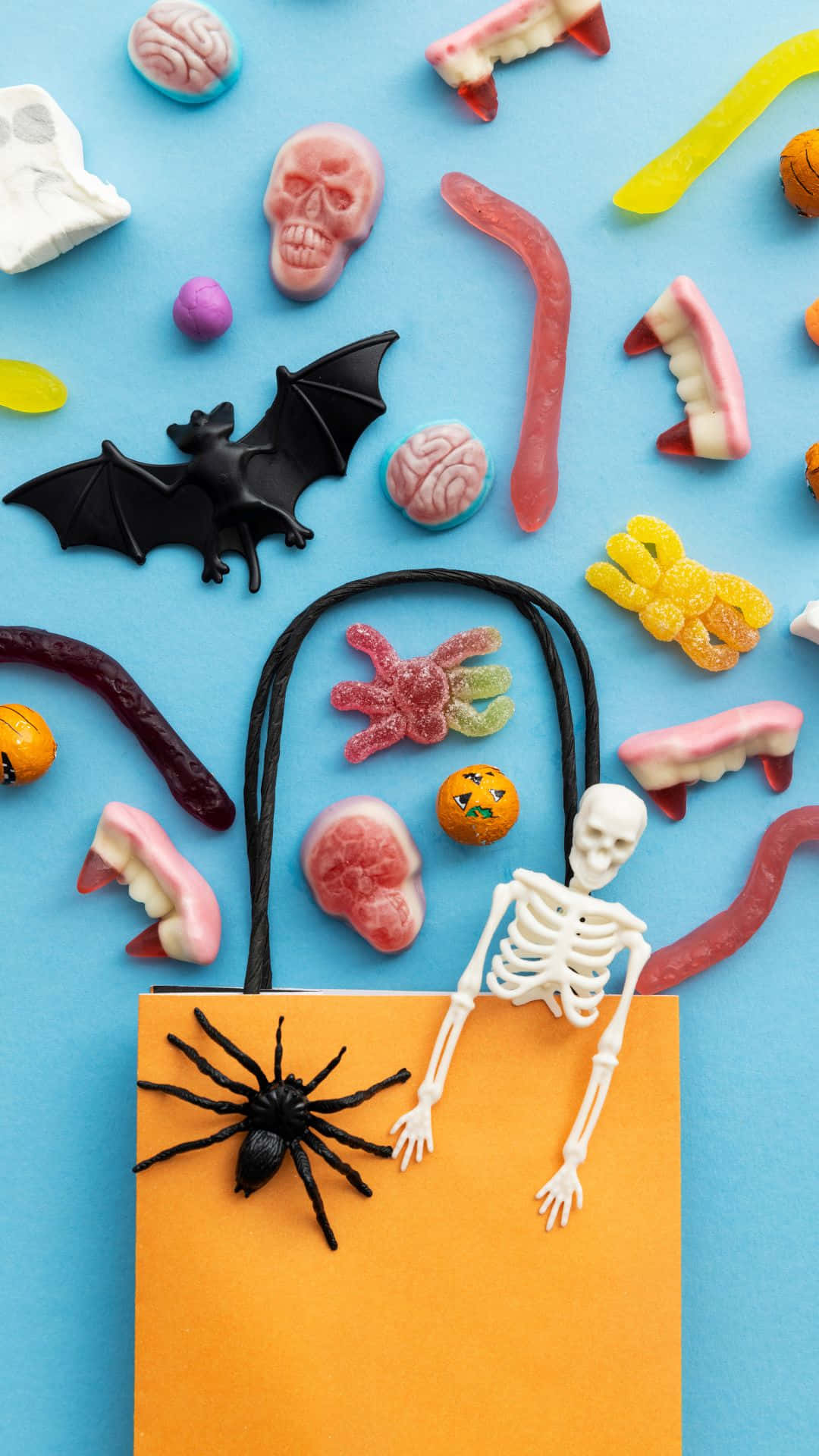 Trick Or Treat Bags 桌布 壁紙 — 萬聖節糖果和玩具的平鋪佈局，包括頭骨、蝙蝠和蜘蛛，圍繞一個橙色袋子排列，袋子裡有一個骷髏探出頭來。