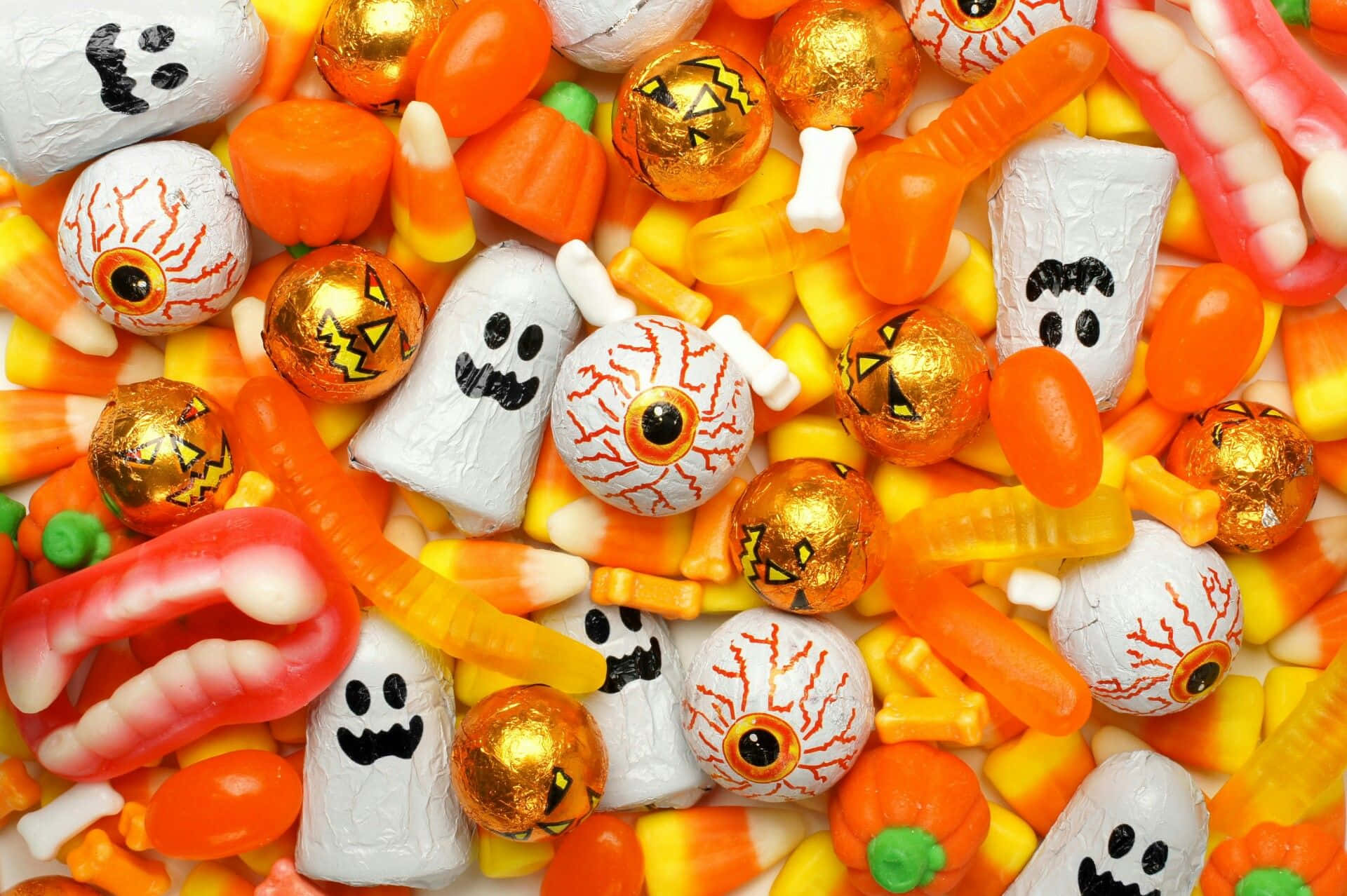 Halloween Treats 桌布 壁紙 — 萬聖節糖果的特寫，包括眼球、幽靈和南瓜形狀的零食，顏色有橙色、黃色和白色。